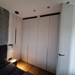 Ferbel - Sypialnia z zabudową szafy wnękowej z białymi frontami bez uchwytów, drewnianą podłogą i tapetą w geometryczny wzór za wezgłowiem łóżka, oświetlenie punktowe i lampka nocna.