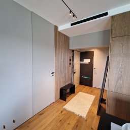 Ferbel - Nowoczesne wnętrze przedpokoju z drewnianą podłogą, szarą szafą w zabudowie, drewnianą ścianą dekoracyjną, czarną ławeczką i jasnym dywanem. Widoczne oświetlenie punktowe na szynie.