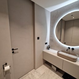 Ferbel - Nowoczesna łazienka w odcieniach szarości z okrągłym lustrem podświetlanym LED, minimalistyczną umywalką nablatową i czarną armaturą, widoczny fragment toalety z czarnym uchwytem na papier toaletowy.