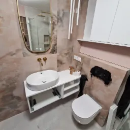 Nowoczesna łazienka z umywalką nablatową, złotą armaturą i marmurkowymi płytkami, widoczne akcesoria i elementy instalacji.