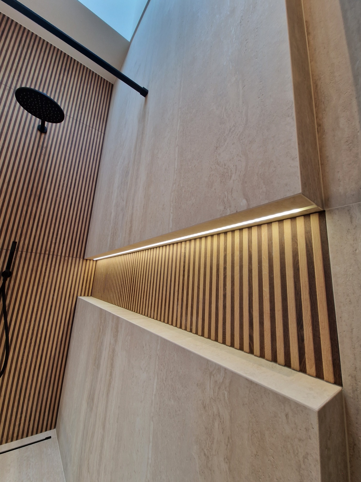 Nowoczesna kabina prysznicowa z drewnianymi panelami, oświetleniem LED i deszczownicą w kolorze czarnym, wykończona płytkami w odcieniach beżu.