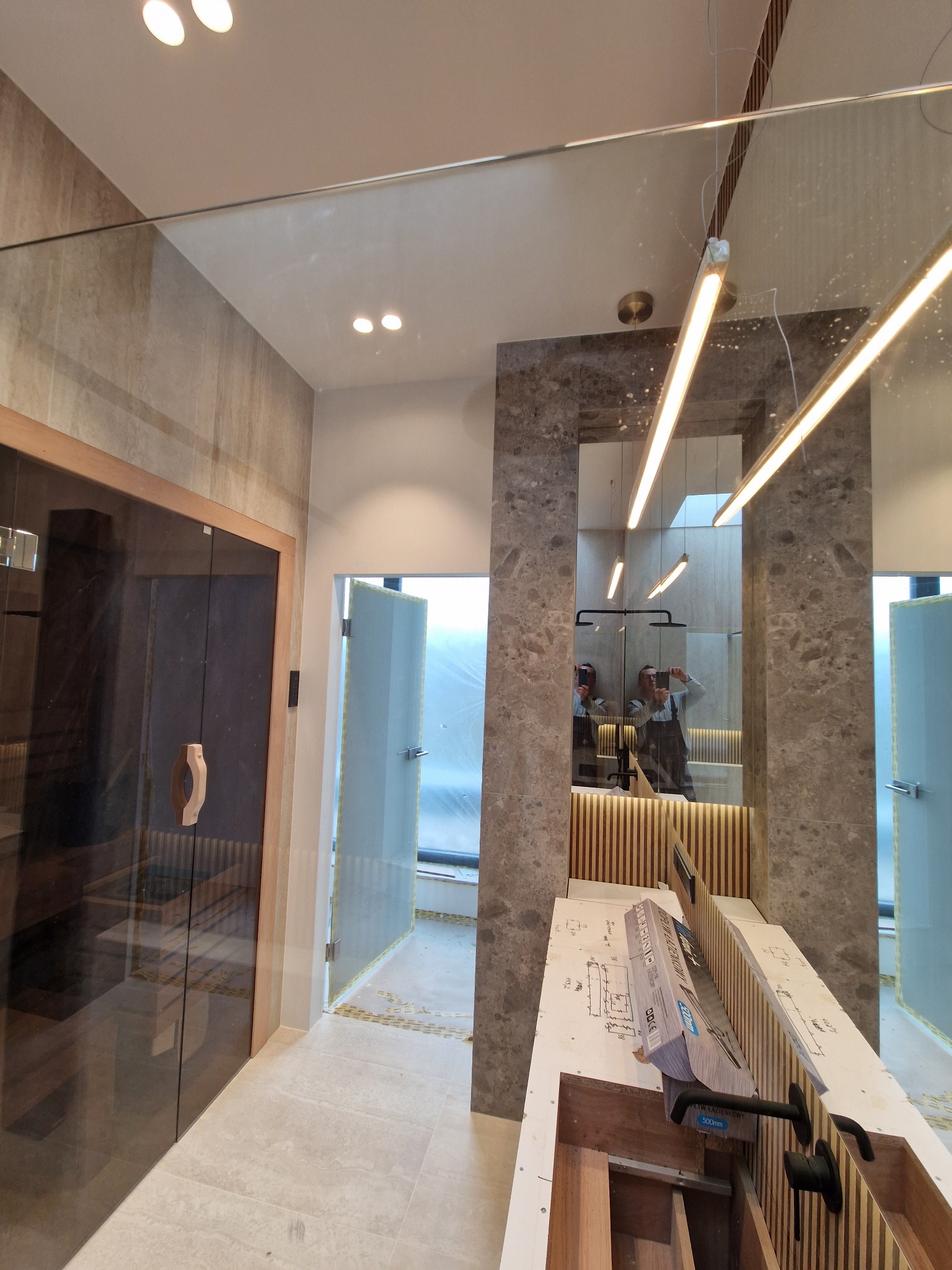 Wnętrze łazienki w trakcie remontu: widoczna sauna z ciemnego drewna, szklana kabina prysznicowa, marmurowe wykończenie ściany z lustrem, oraz przygotowane elementy do montażu armatury łazienkowej.