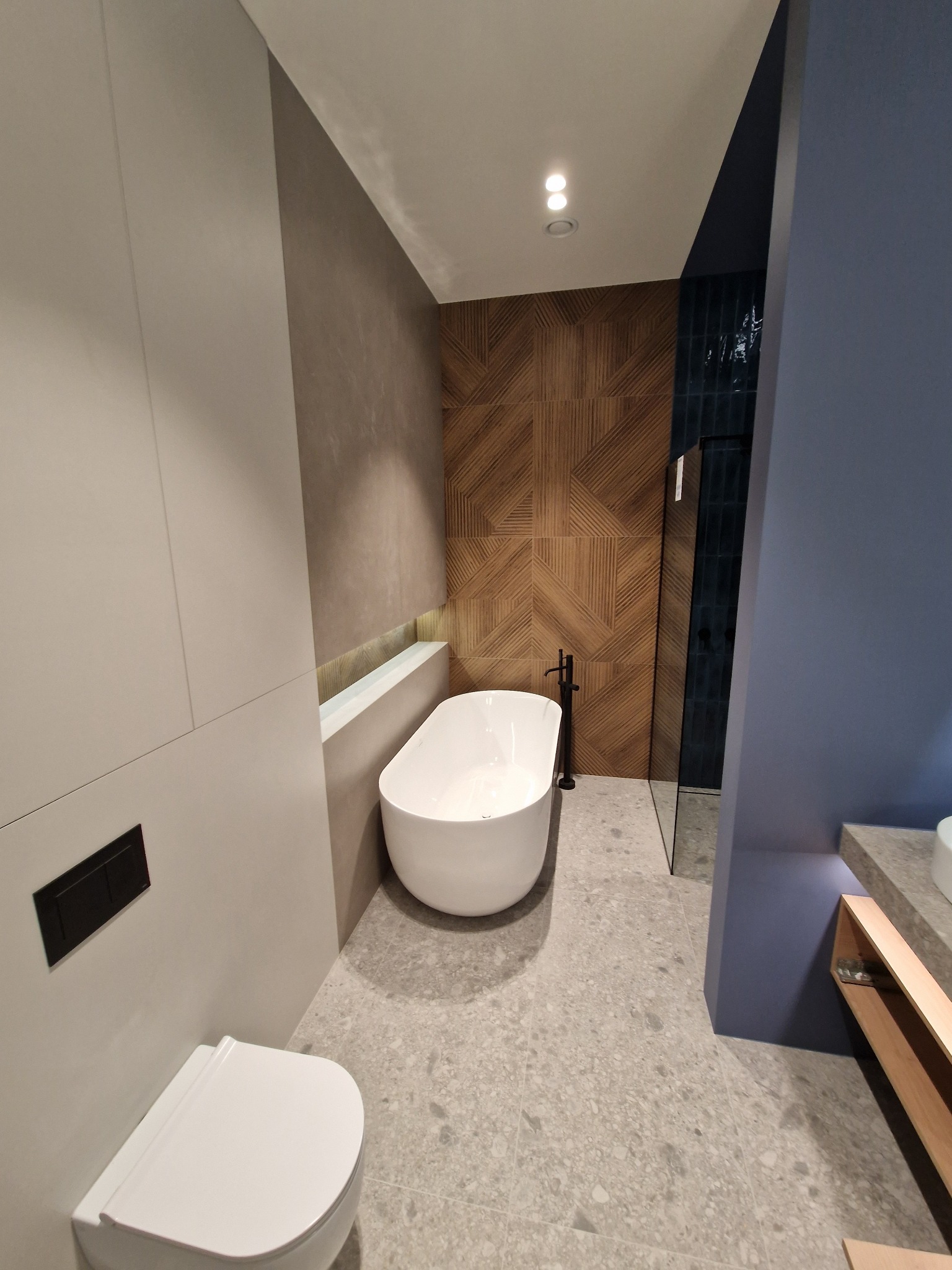 Nowoczesna łazienka z wolnostojącą białą wanną, drewnianą ścianą z geometrycznym wzorem, szarą podłogą z płytek imitujących kamień i podwieszaną toaletą z czarnym przyciskiem.