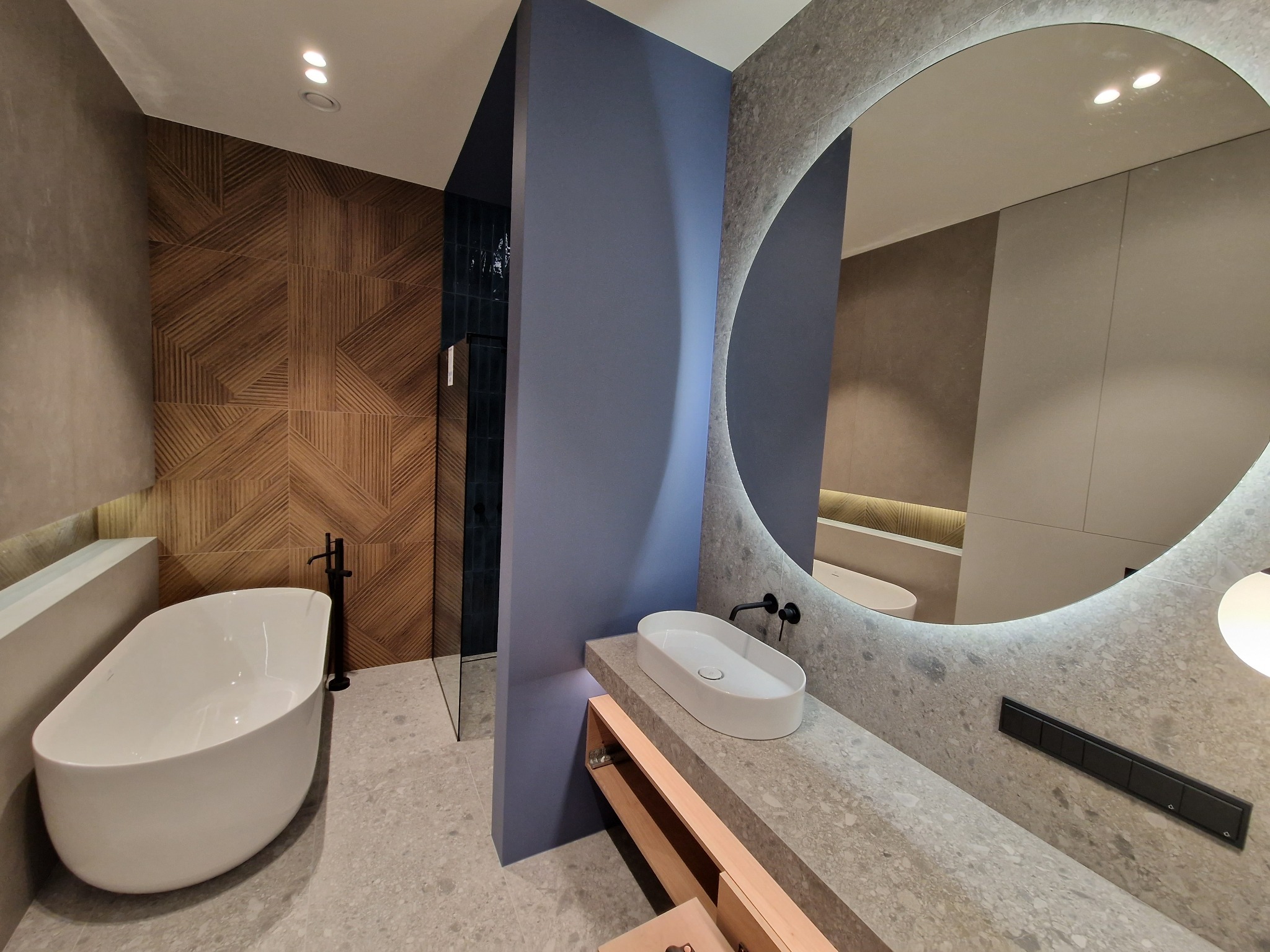 Nowoczesna łazienka z wolnostojącą białą wanną, umywalką nablatową na blacie z szarego kamienia, okrągłym lustrem z podświetleniem LED i ścianą z drewnianej mozaiki w geometryczny wzór.