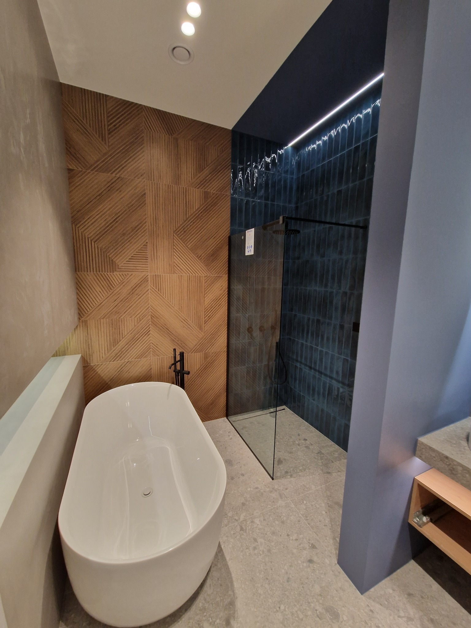 Nowoczesna łazienka z wolnostojącą wanną, kabiną prysznicową z ciemnymi płytkami i drewnianą ścianą z geometrycznym wzorem, oświetleniem LED i szarą podłogą.