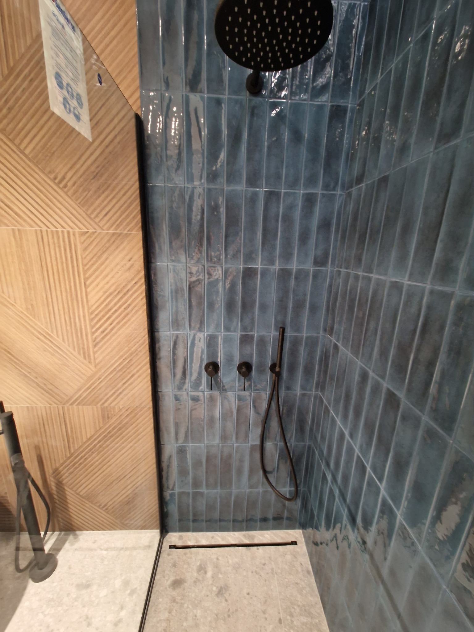 Nowoczesna kabina prysznicowa z turkusowymi, błyszczącymi płytkami ułożonymi pionowo, z czarną deszczownicą i armaturą, oraz panel dekoracyjny z drewnopodobnych płytek z geometrycznym wzorem.