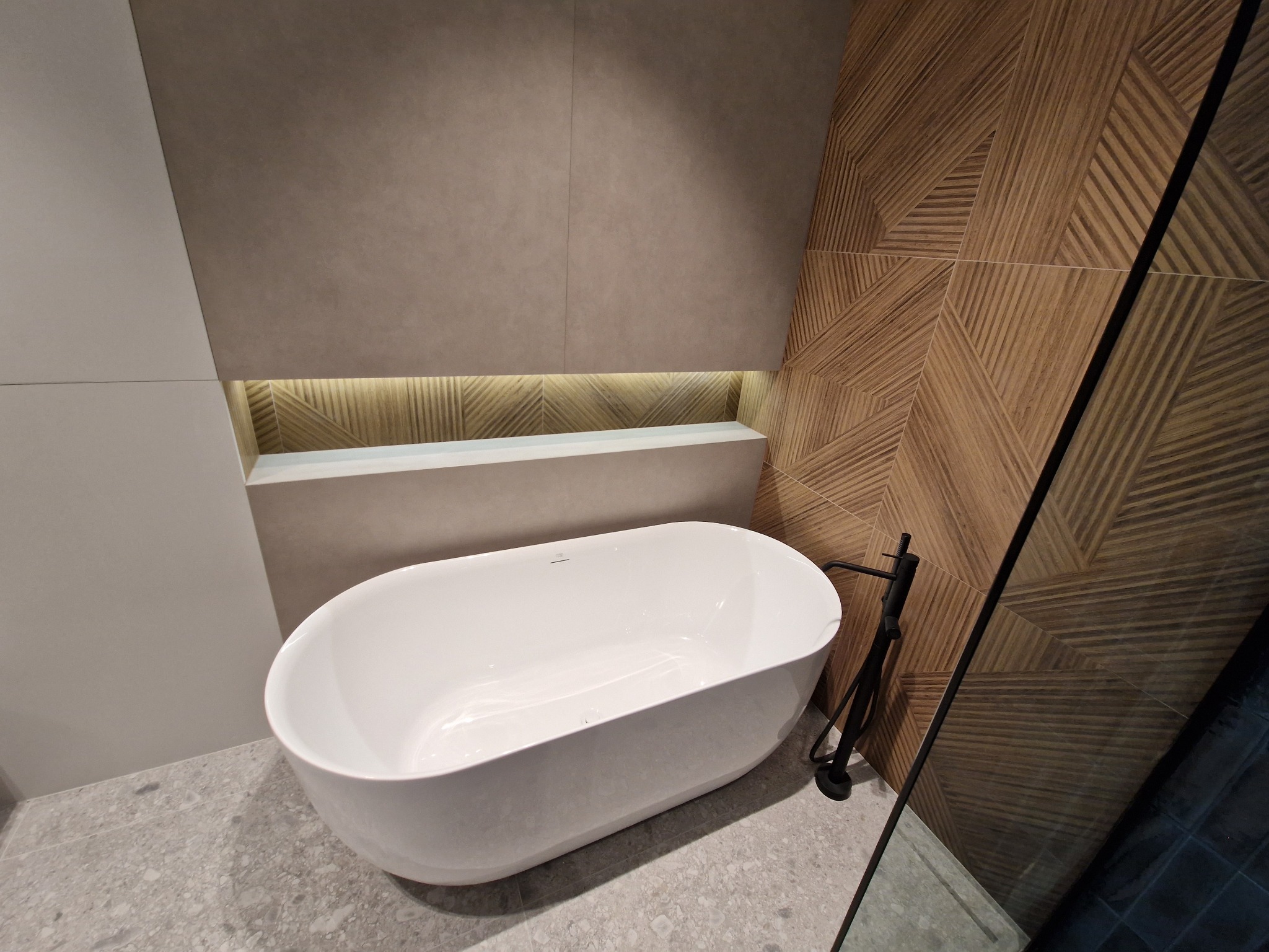 Wolnostojąca biała wanna z czarną armaturą na tle ściany wyłożonej płytkami w geometryczny wzór drewna, oświetlona półka w zabudowie.