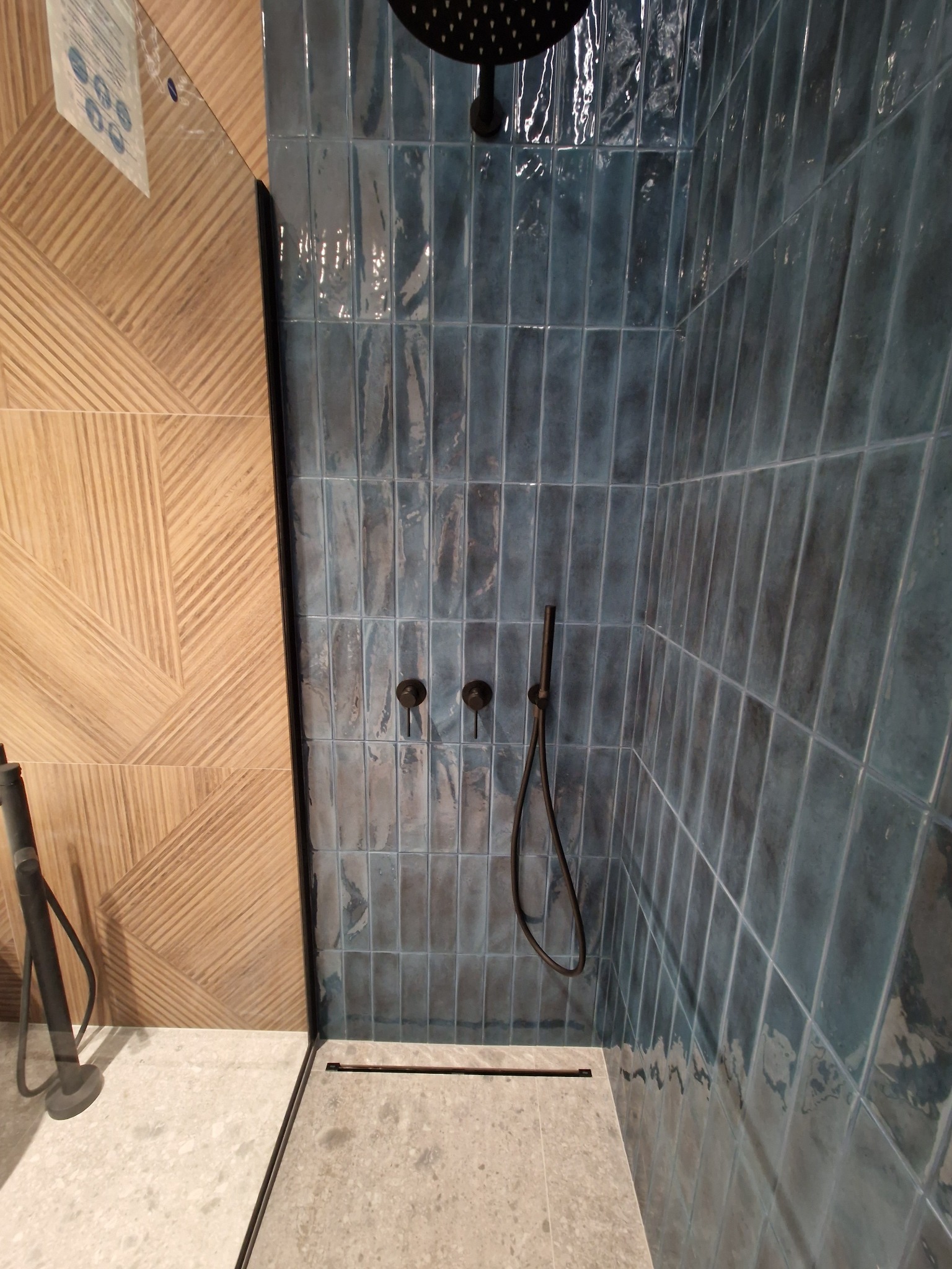 Nowoczesny prysznic z niebieskimi, błyszczącymi płytkami wertykalnymi i drewnopodobną ścianą z geometrycznym wzorem, czarna armatura i odpływ liniowy.