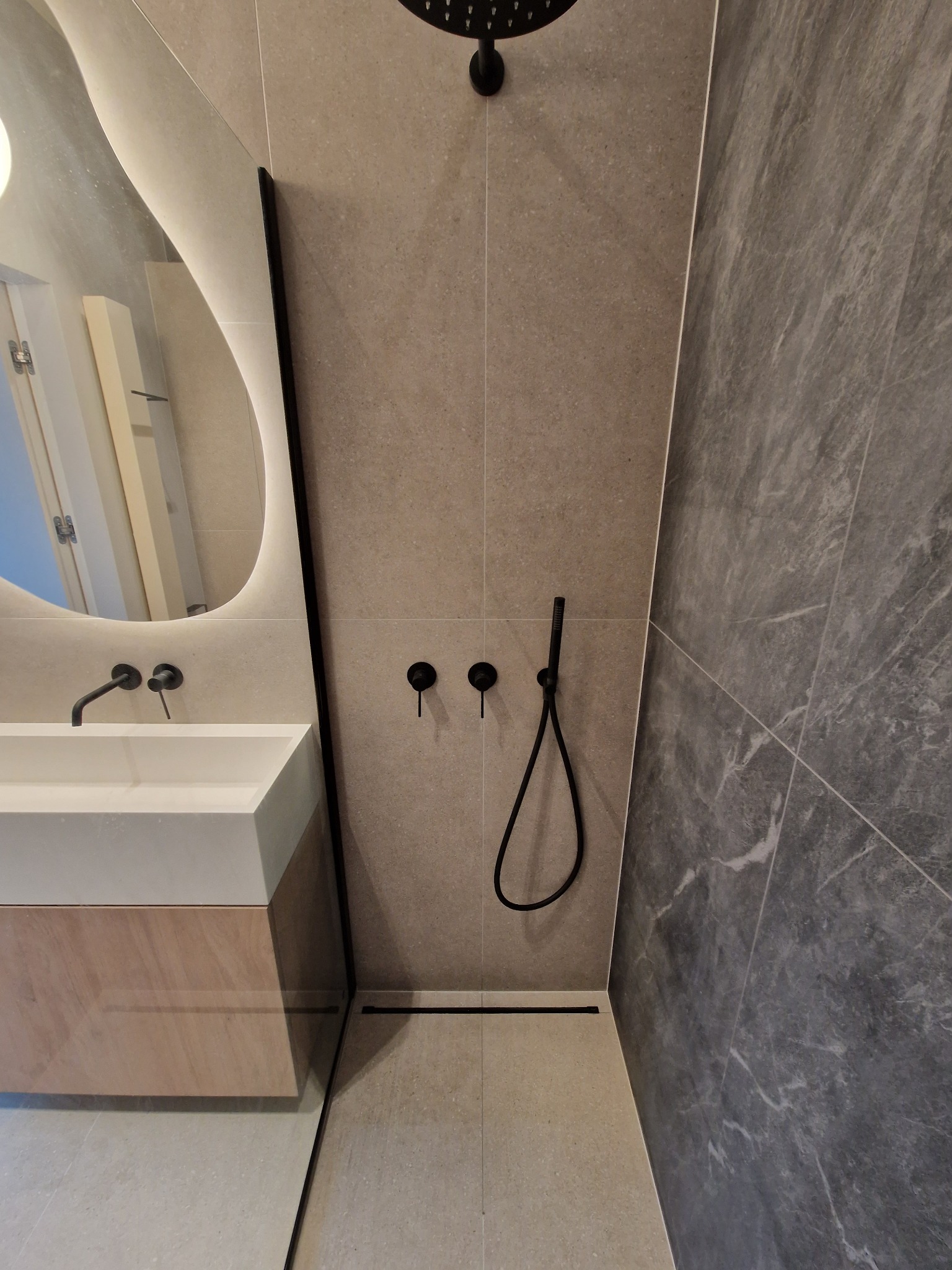 Nowoczesna łazienka z deszczownicą, czarną armaturą i asymetrycznym lustrem z podświetleniem LED.