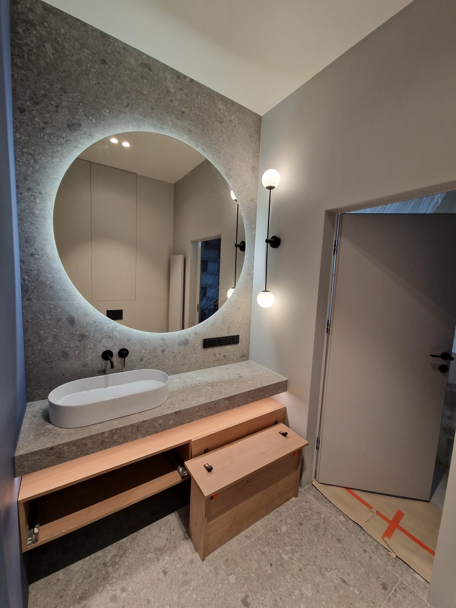 Nowoczesna łazienka z owalną umywalką nablatową, podświetlanym okrągłym lustrem i szafkami z jasnego drewna; zabezpieczenie podłogi kartonem z taśmą w pobliżu drzwi.