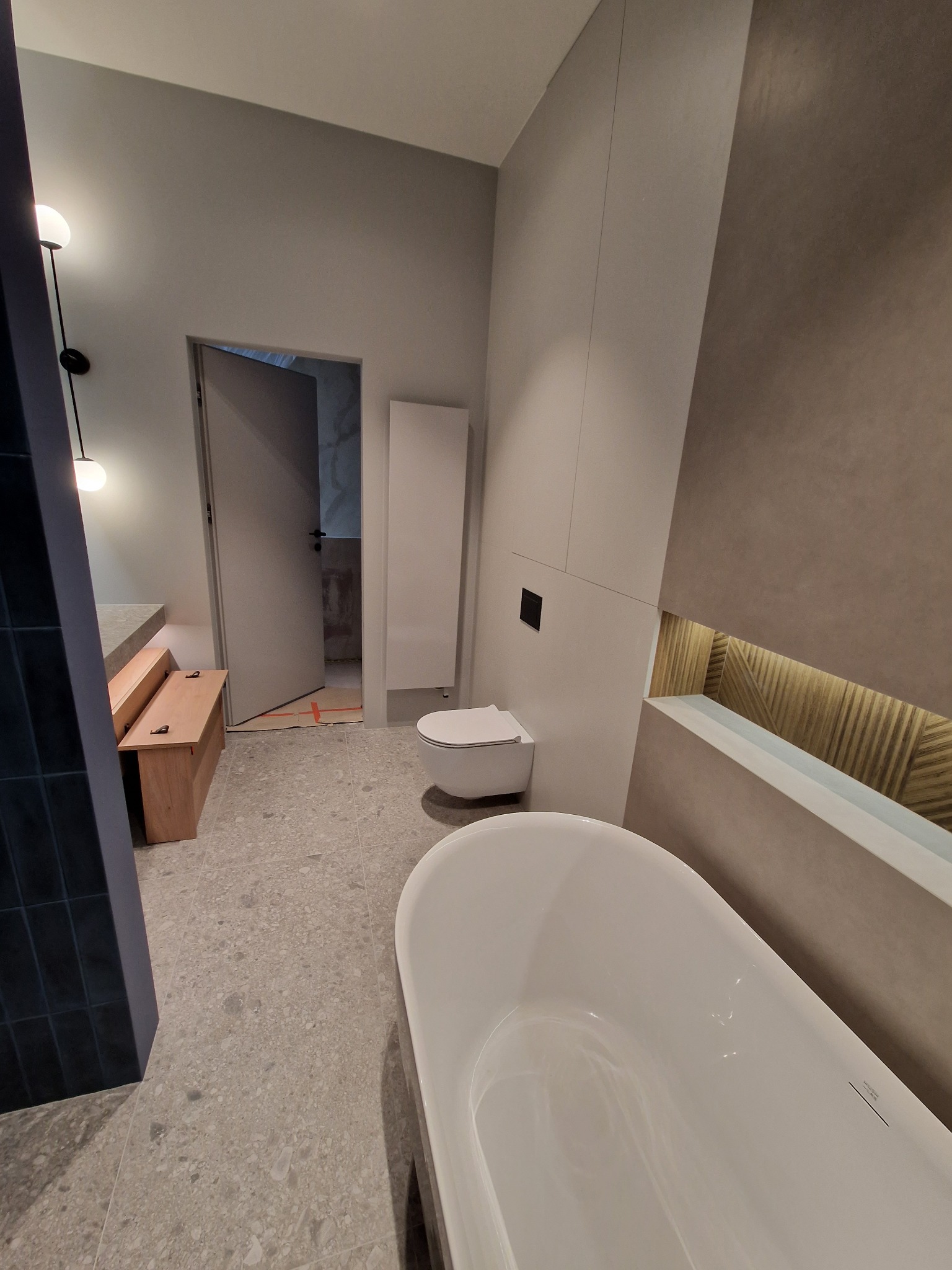 Nowoczesna łazienka z wolnostojącą białą wanną, toaletą w zabudowie i drewnianym akcentem w ścianie, szara podłoga z wzorem.