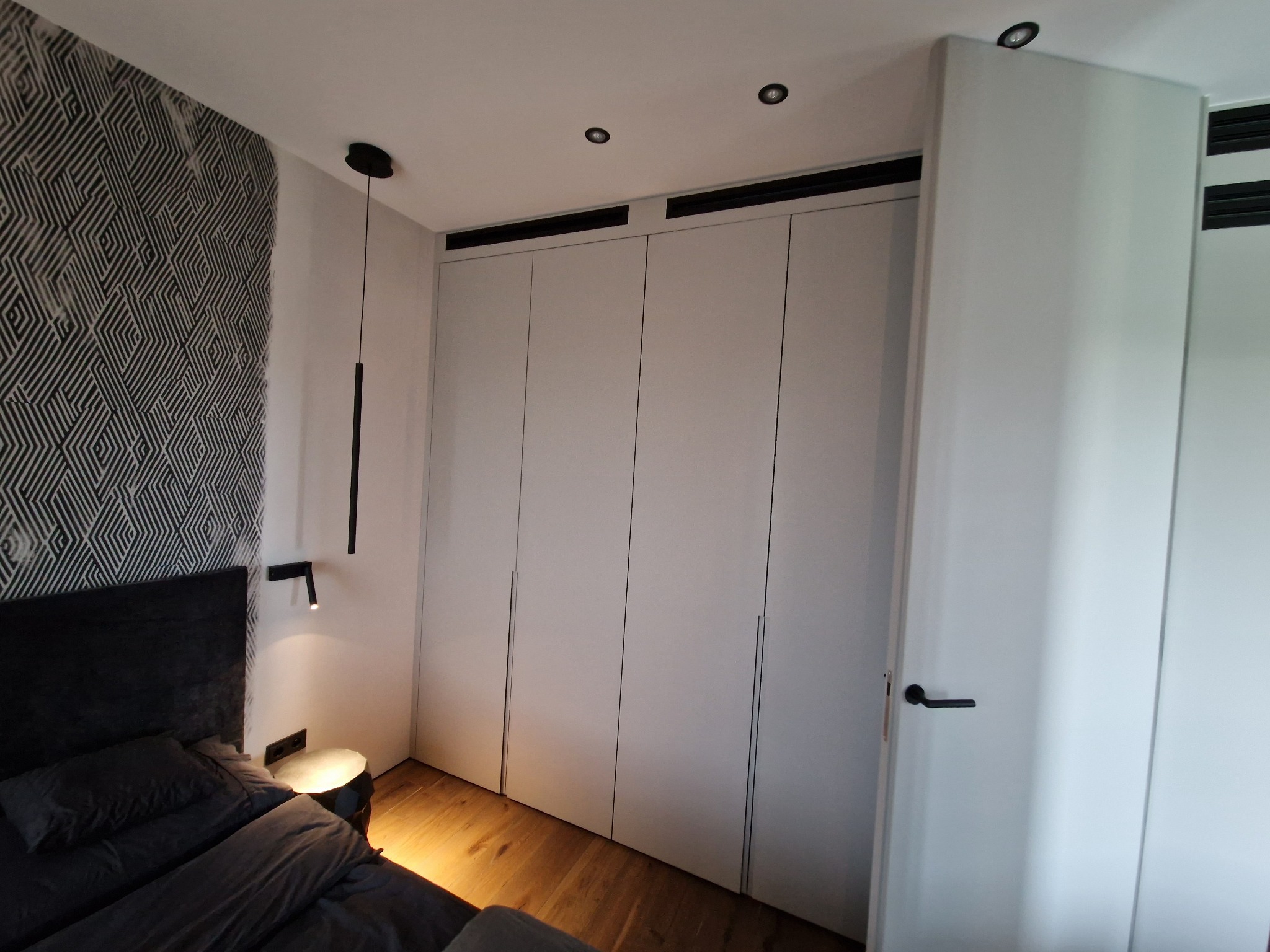 Sypialnia z zabudową szafy wnękowej z białymi frontami bez uchwytów, drewnianą podłogą i tapetą w geometryczny wzór za wezgłowiem łóżka, oświetlenie punktowe i lampka nocna.