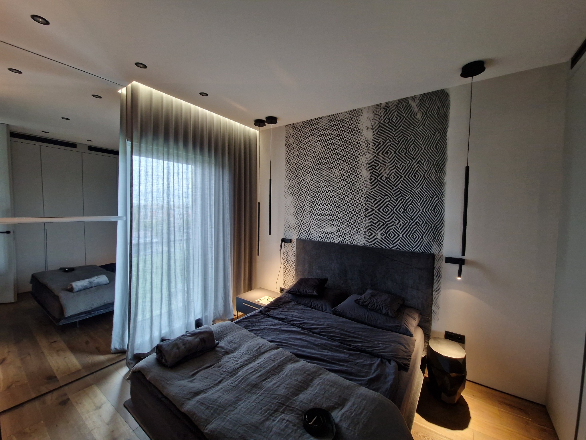 Nowoczesna sypialnia z łóżkiem w odcieniach szarości, geometryczną tapetą za wezgłowiem i odbiciem pokoju w lustrzanej ścianie, z minimalistycznym oświetleniem punktowym i zasłonami.
