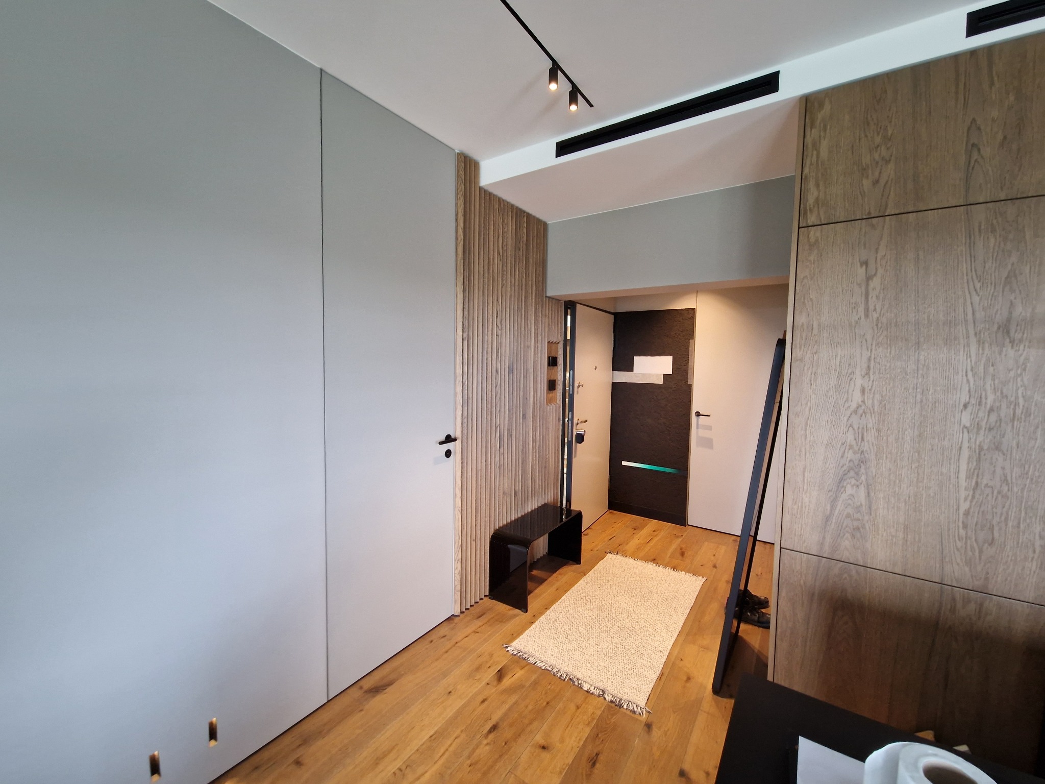 Nowoczesne wnętrze przedpokoju z drewnianą podłogą, szarą szafą w zabudowie, drewnianą ścianą dekoracyjną, czarną ławeczką i jasnym dywanem. Widoczne oświetlenie punktowe na szynie.