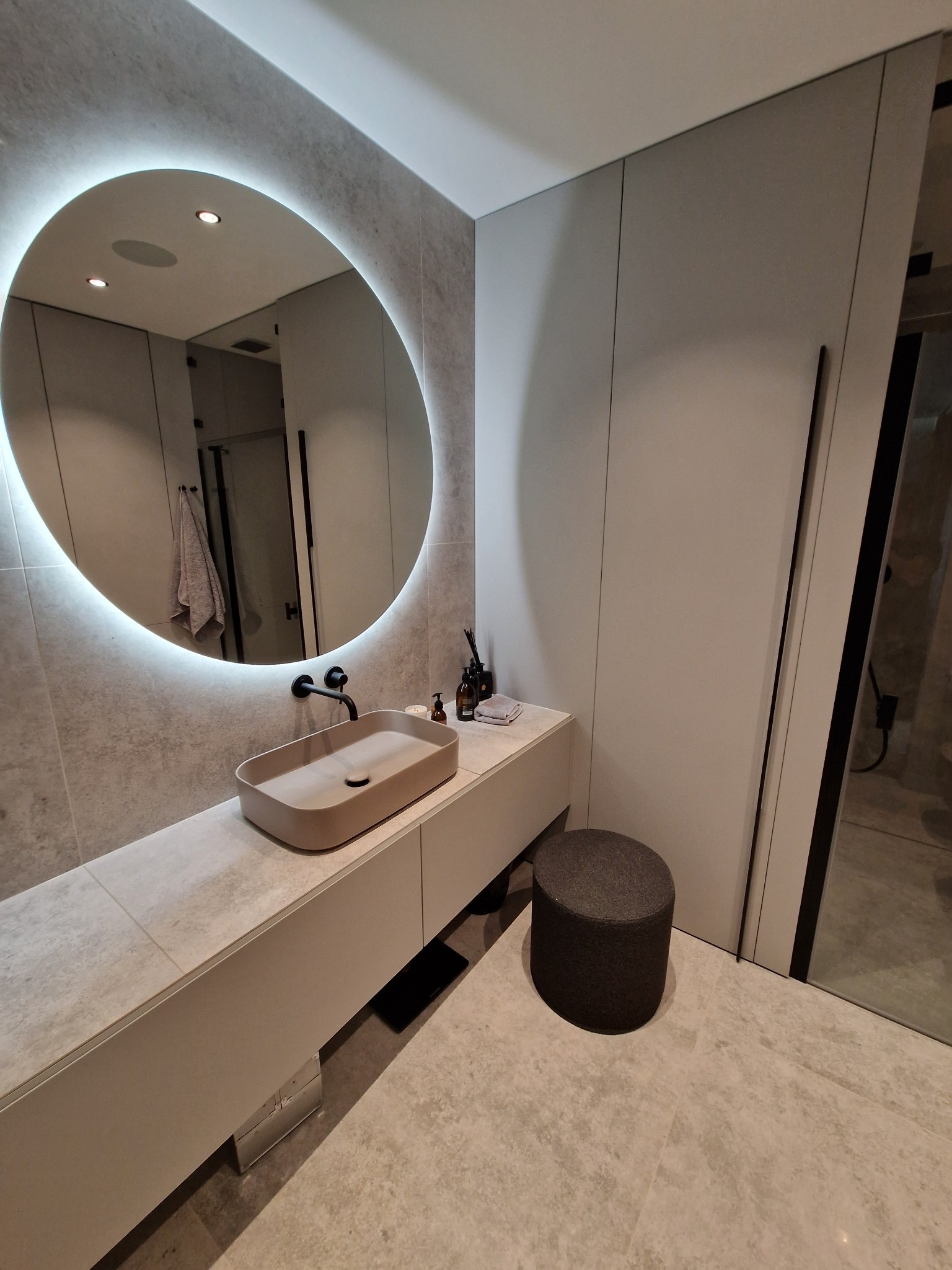 Nowoczesna łazienka z owalnym lustrem podświetlanym LED, beżową umywalką nablatową i szarą mozaiką na ścianie oraz ciemnoszarym stołkiem obok szafki umywalkowej.
