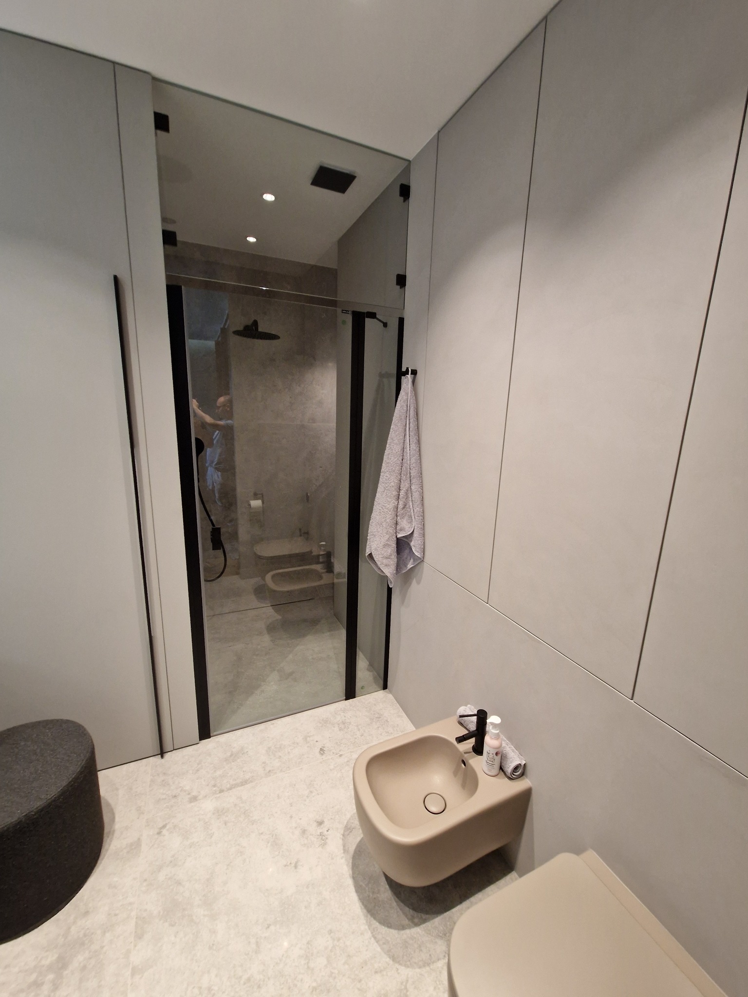 Nowoczesna łazienka z beżową armaturą, szklanym prysznicem z czarną ramą, szarą ścianą z panelami i okrągłym pufem, widoczna osoba w odbiciu.