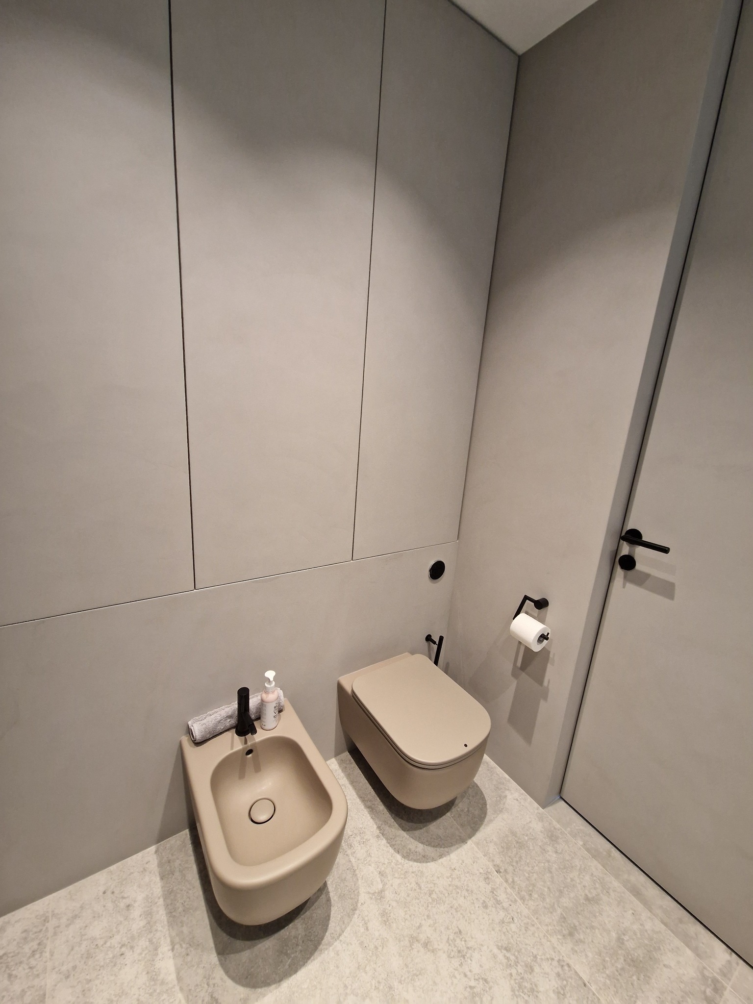 Minimalistyczna łazienka w odcieniach szarości z beżową armaturą: wisząca toaleta i bidet z czarną baterią, na blacie bidetu ręcznik i dozownik mydła.