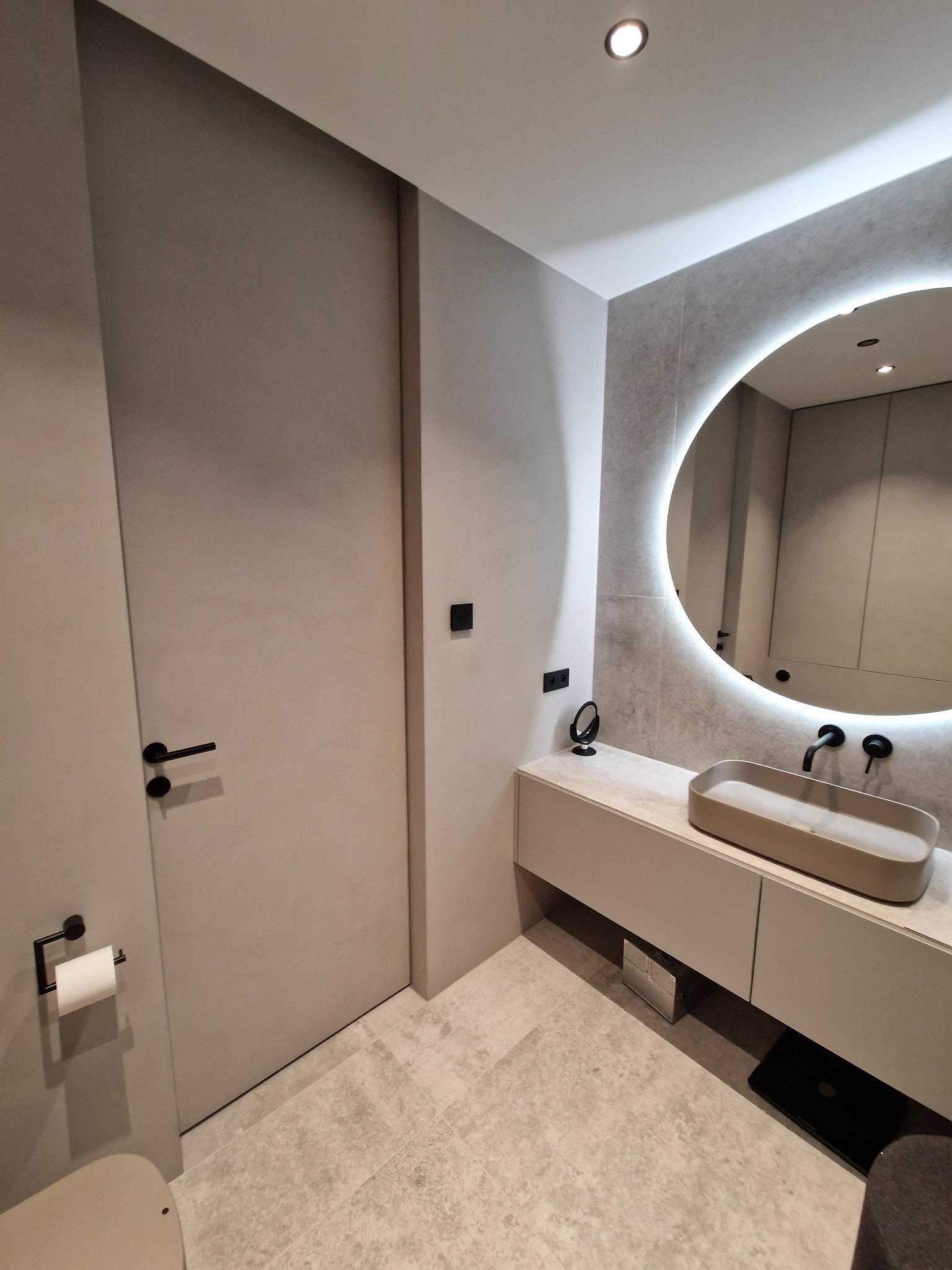 Nowoczesna łazienka w odcieniach szarości z okrągłym lustrem podświetlanym LED, minimalistyczną umywalką nablatową i czarną armaturą, widoczny fragment toalety z czarnym uchwytem na papier toaletowy.