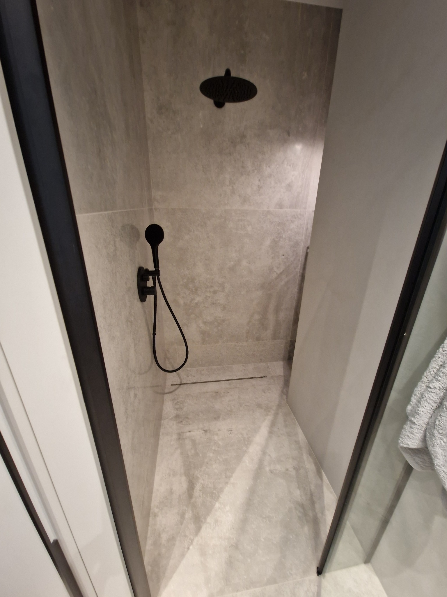 Nowoczesna kabina prysznicowa z szarymi płytkami imitującymi marmur, czarną armaturą i odpływem liniowym w podłodze.