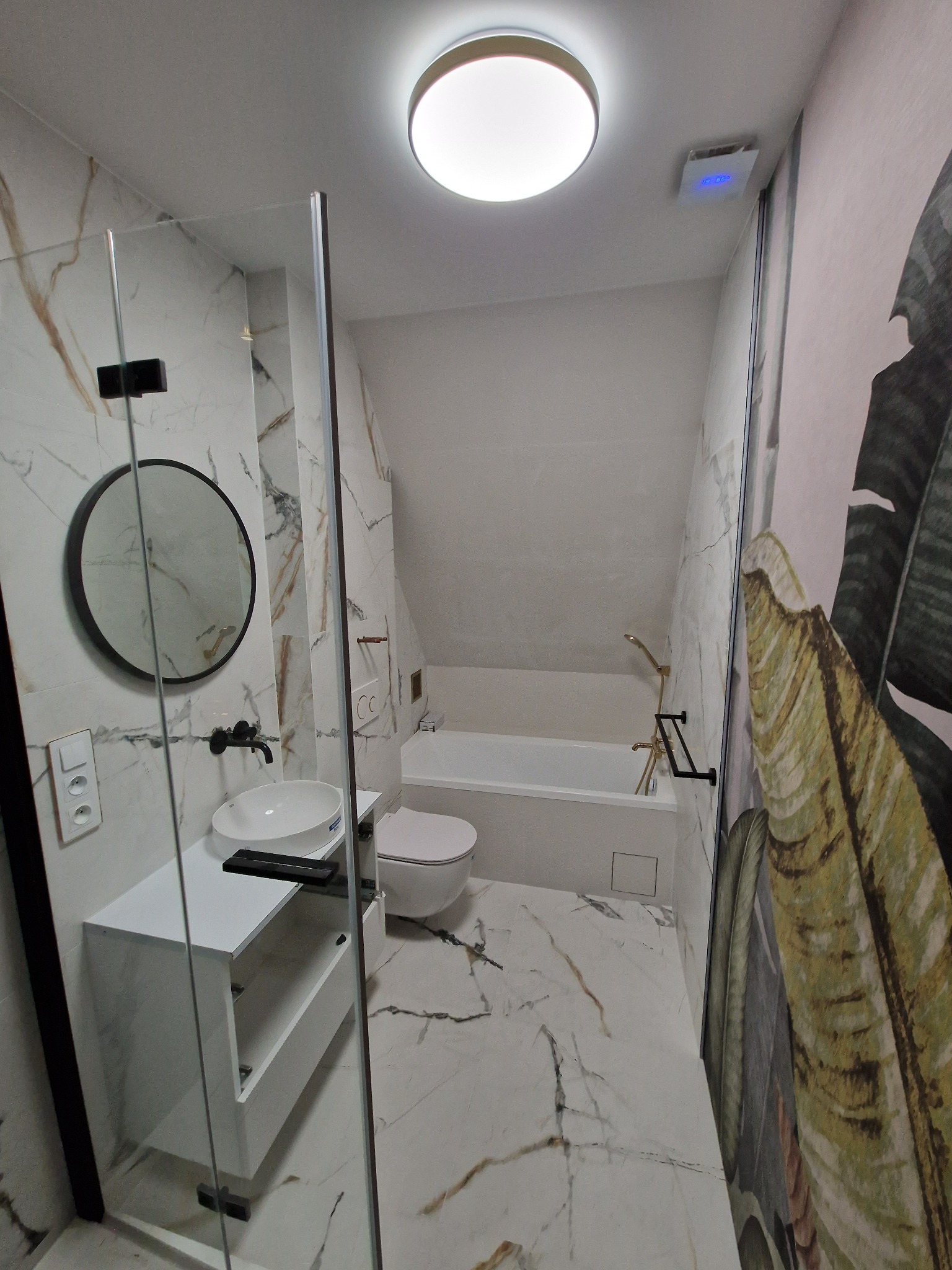 Nowoczesna łazienka w odcieniach bieli i złota z marmurowymi płytkami, czarną armaturą i okrągłym lustrem, kabina prysznicowa typu walk-in, dekoracyjna tapeta z motywem roślinnym na ścianie.