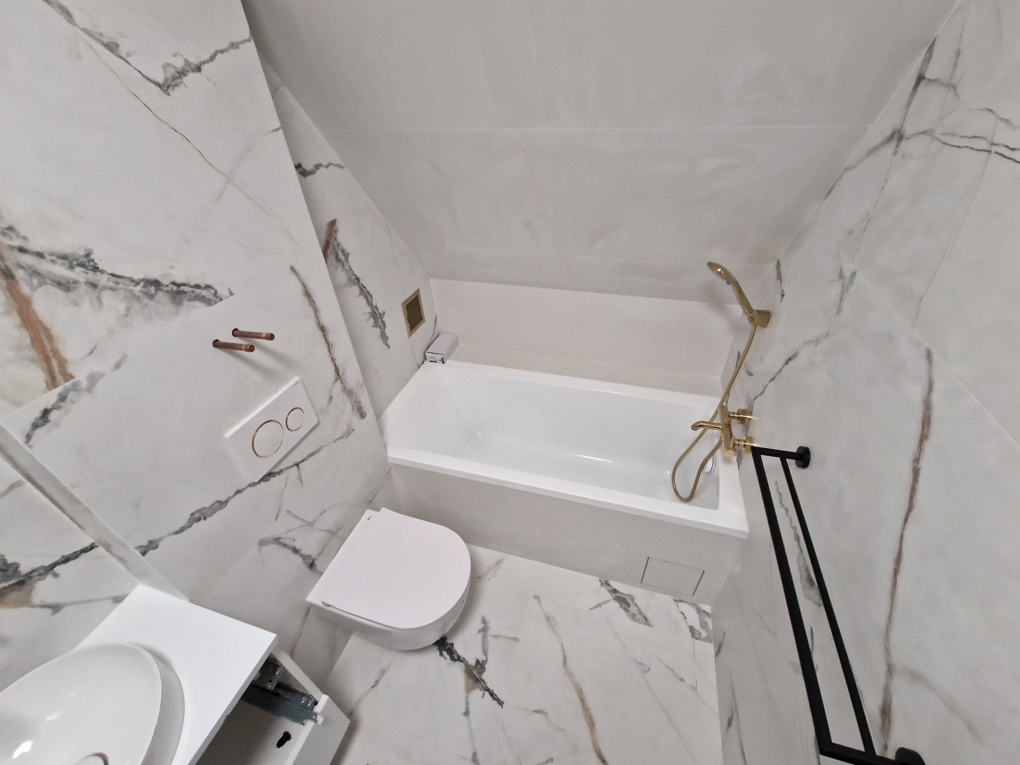 Wykończona łazienka na poddaszu w bieli i imitacji marmuru, z wanną, podwieszaną toaletą i złotą armaturą oraz czarnym grzejnikiem drabinkowym.