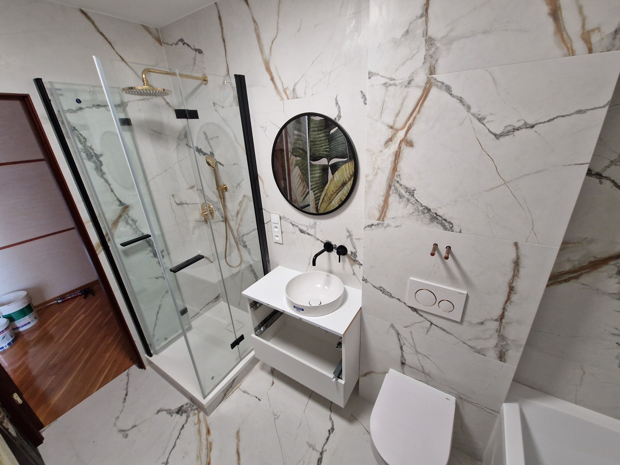 Nowoczesna łazienka z kabiną prysznicową typu walk-in z czarnymi profilami, okrągłym lustrem z motywem roślinnym, białą umywalką nablatową i płytkami imitującymi marmur.