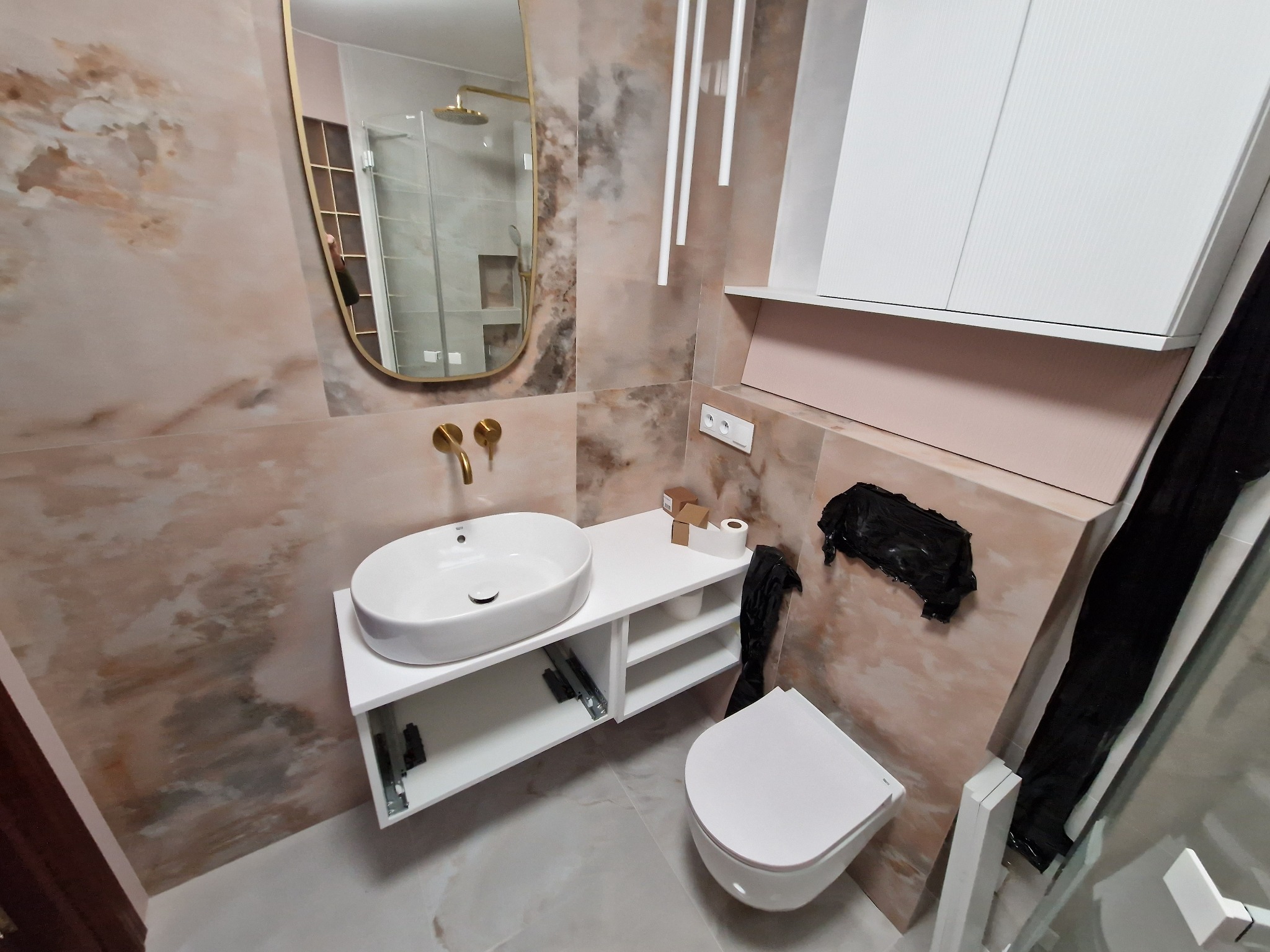 Nowoczesna łazienka z umywalką nablatową, złotą armaturą i marmurkowymi płytkami, widoczne akcesoria i elementy instalacji.