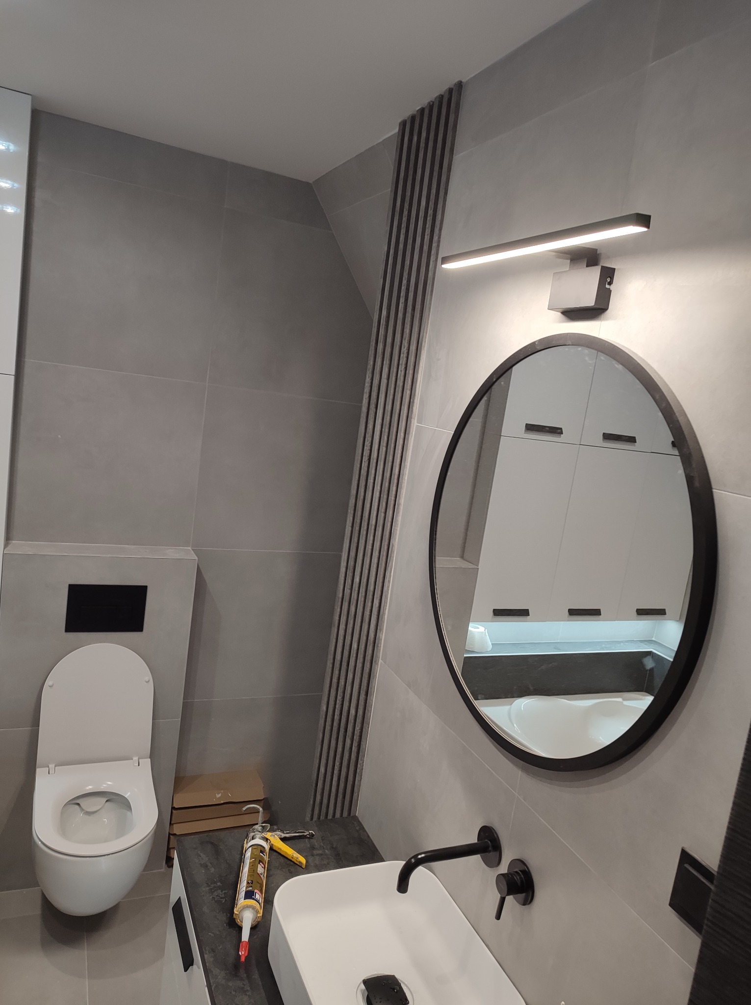 Nowoczesna łazienka w szarościach z okrągłym lustrem podświetlanym lampą LED, wiszącą toaletą i umywalką, widoczna tuba z silikonem i narzędzia na blacie.