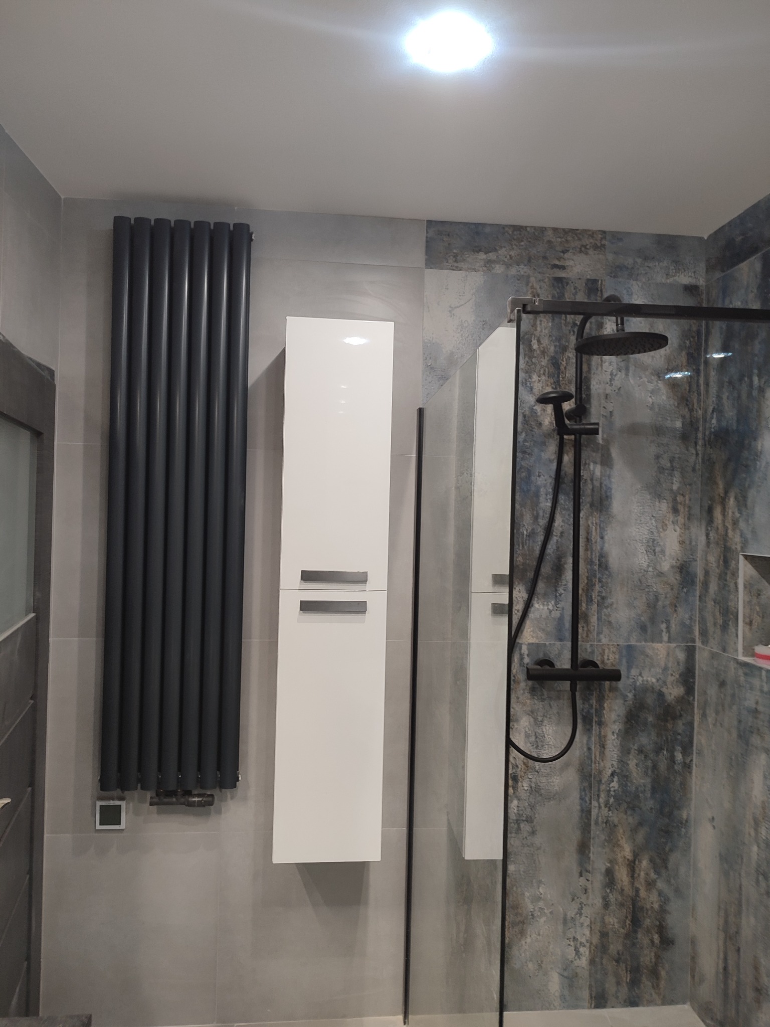 Nowoczesna łazienka z kabiną prysznicową typu walk-in, grzejnikiem żeberkowym w kolorze antracytowym, wiszącą białą szafką i płytkami imitującymi marmur w odcieniach szarości i błękitu.