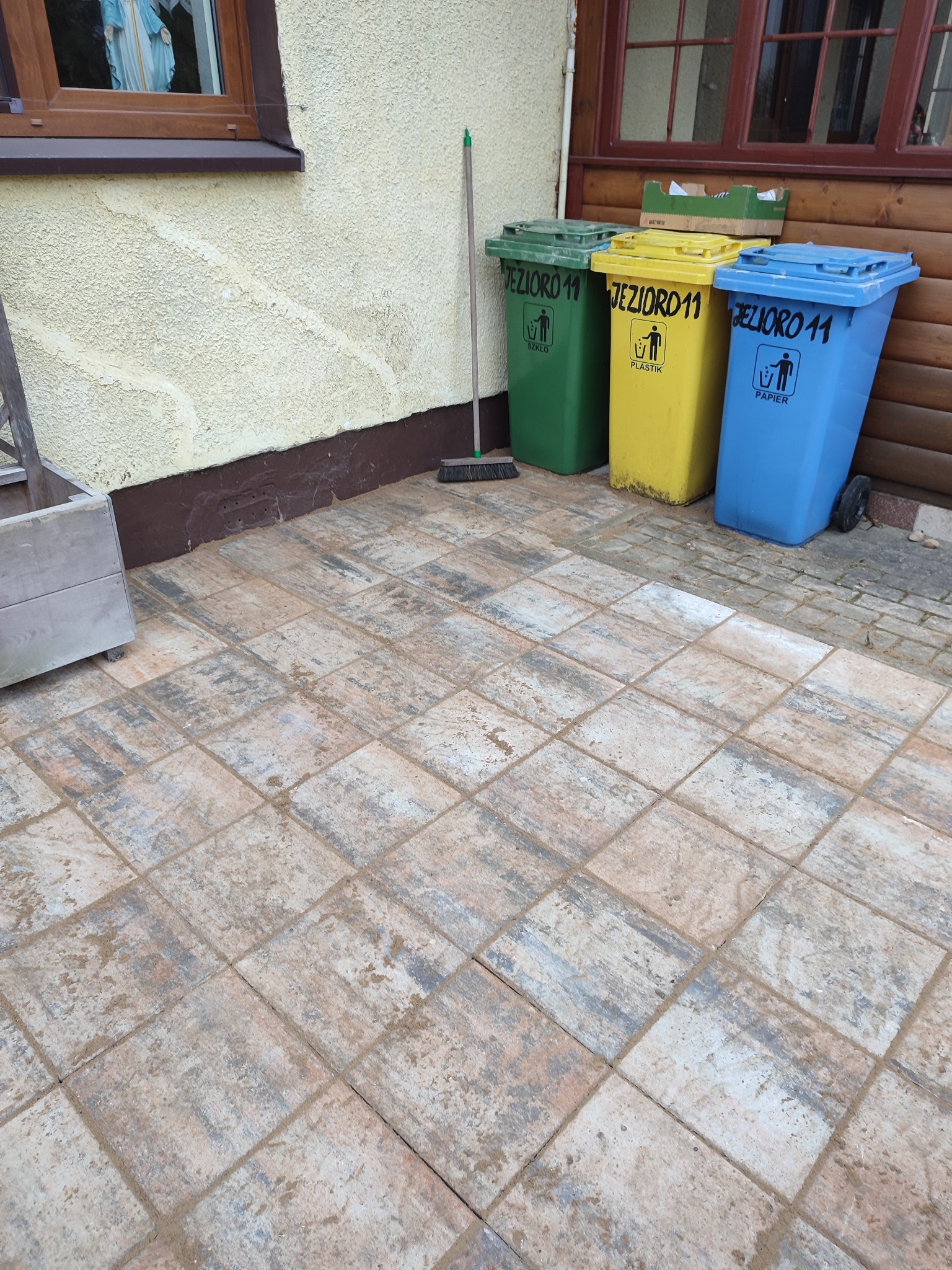 Nowo ułożona kostka brukowa w odcieniach beżu i szarości, z widocznymi trzema pojemnikami na segregację śmieci w tle, oznaczonymi napisami 'Jezioro 11' i symbolami szkła, plastiku i papieru.