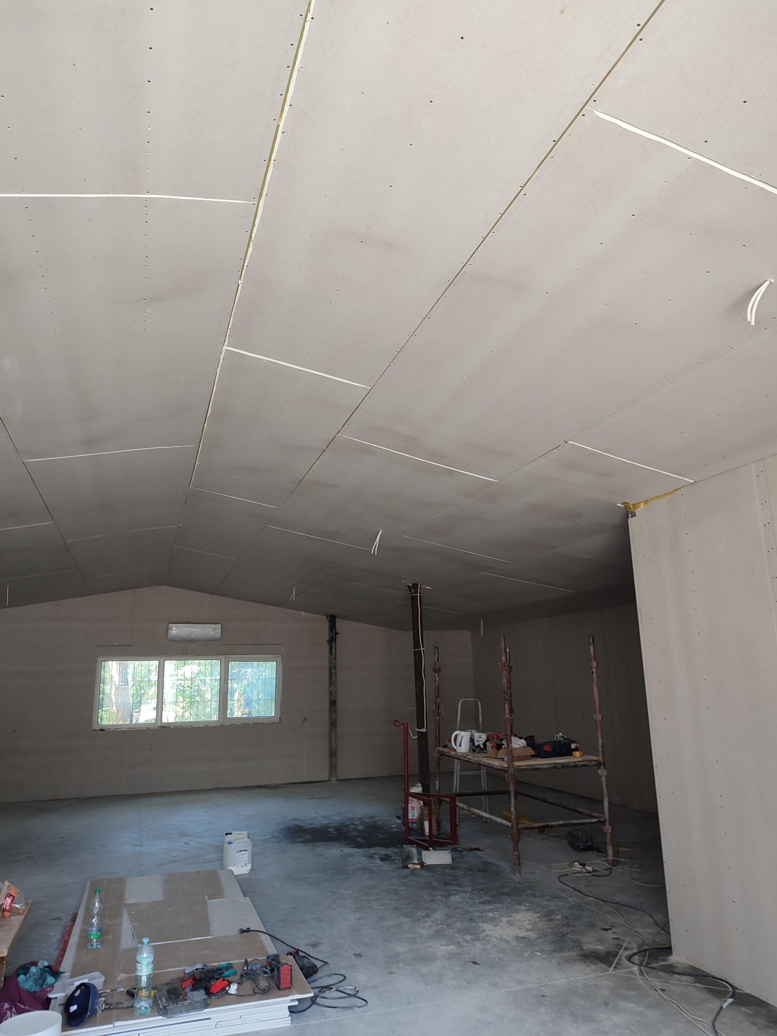 Montaż płyt kartonowo-gipsowych na suficie podwieszanym w surowym wnętrzu z widocznymi elementami konstrukcyjnymi i narzędziami pracy na podłodze.