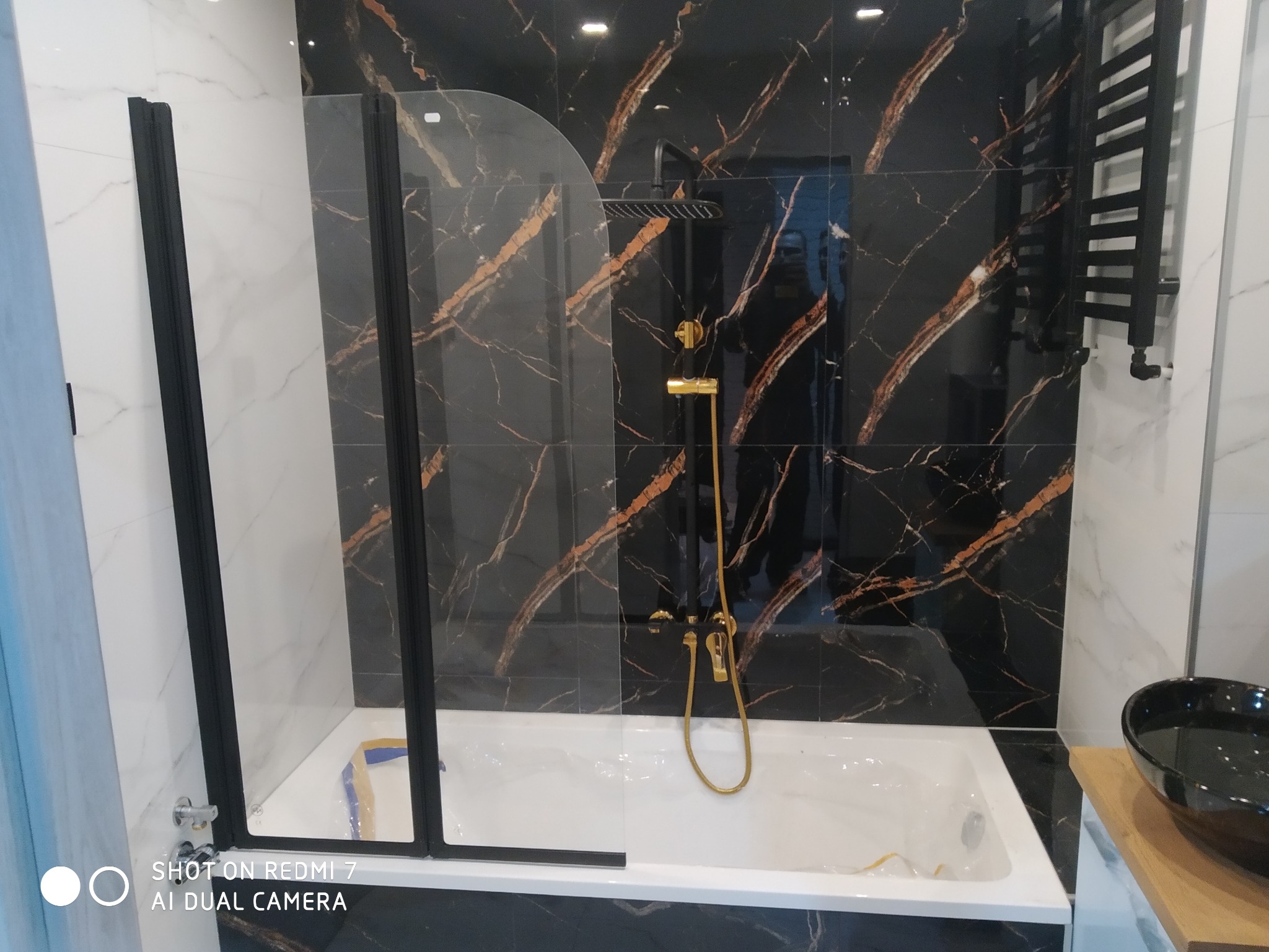 Nowoczesna łazienka z czarno-złotymi płytkami imitującymi marmur, białą wanną i czarną armaturą oraz grzejnikiem drabinkowym, szklany parawan nawannowy z czarnymi profilami.