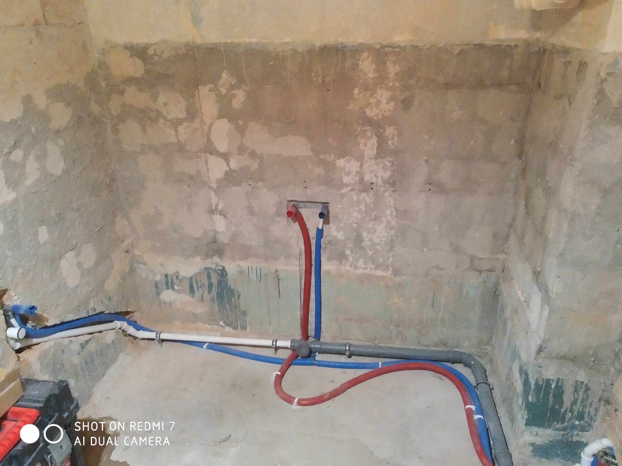 Surowy stan łazienki w trakcie prac hydraulicznych: widoczne rury instalacji wodnej (czerwone i niebieskie) prowadzone wzdłuż ściany, z surowym tynkiem i śladami po kuciu.