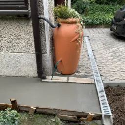 Świeżo wylany beton na tarasie przydomowym, obok ściany budynku z rynną i ozdobną donicą, widoczny drenaż liniowy.