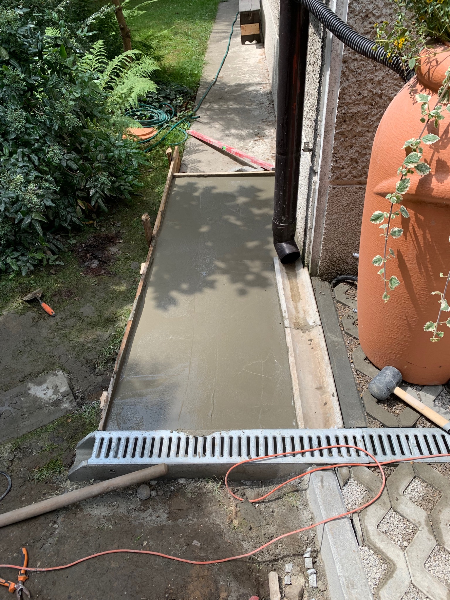 Wylewka betonowa w drewnianym szalunku, obok rynna odwadniająca i zbiornik na deszczówkę z pnączem, widoczne narzędzia budowlane.