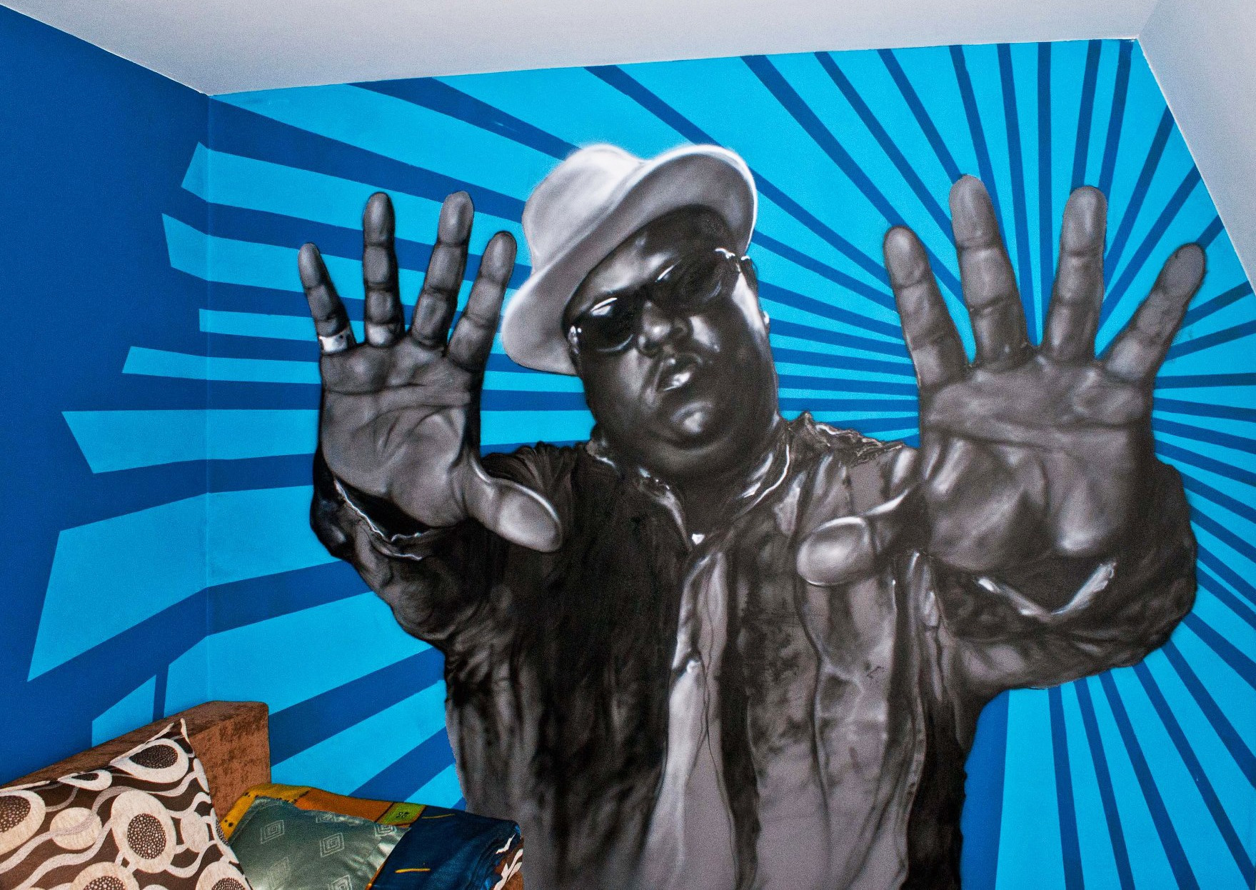 Czarno-biały portret Biggie Smalls w kapeluszu i okularach, namalowany na błękitnej ścianie z promienistym wzorem, widoczny fragment łóżka z poduszkami w dolnej części kadru.