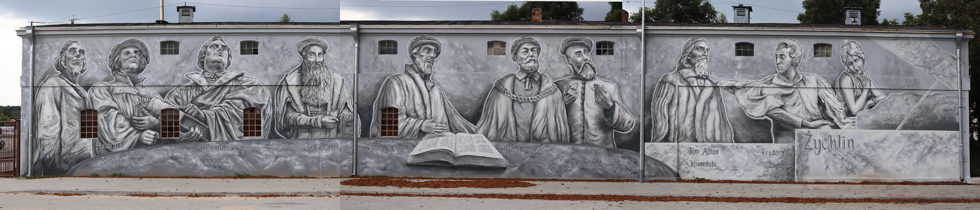Szary mural na ścianie budynku w Żychlinie przedstawiający postacie historyczne i otwartą księgę, z widocznymi oknami w tle i chodnikiem z liśćmi u podstawy ściany.