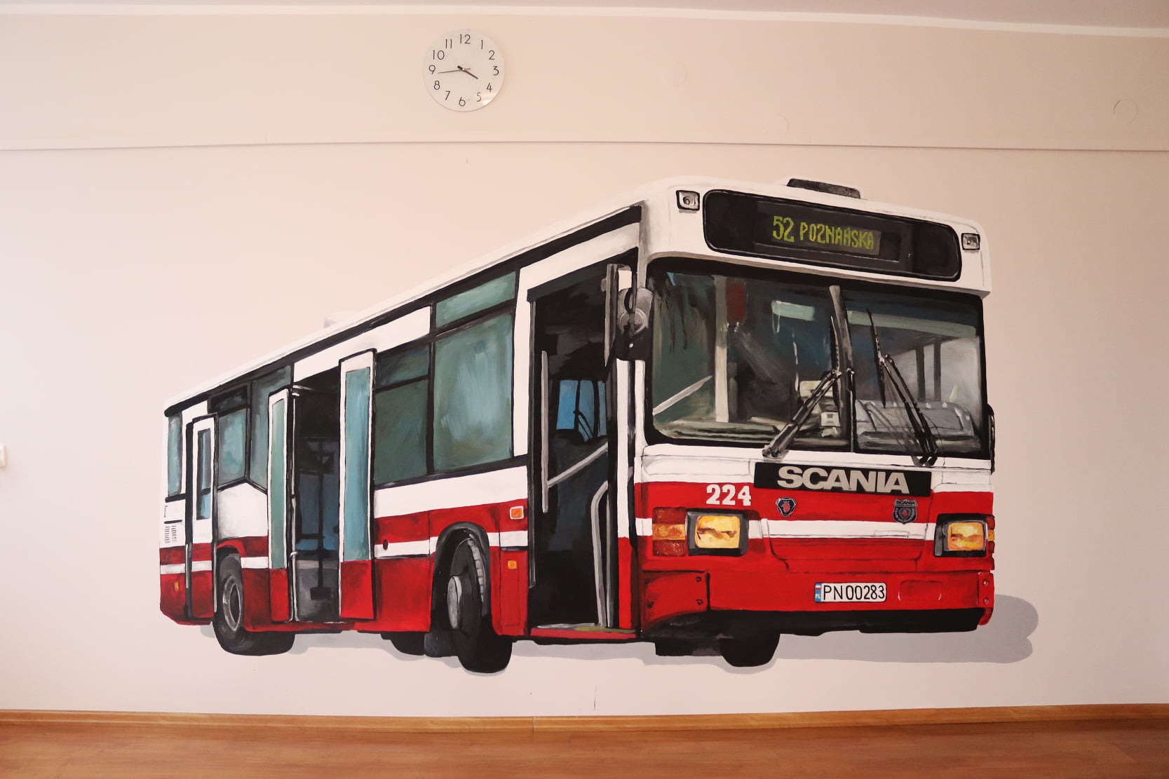 Realistyczne malowidło ścienne przedstawiające czerwono-biały autobus miejski Scania z numerem 52 i tablicą rejestracyjną, widoczne na białej ścianie nad drewnianą podłogą, z okrągłym zegarem...