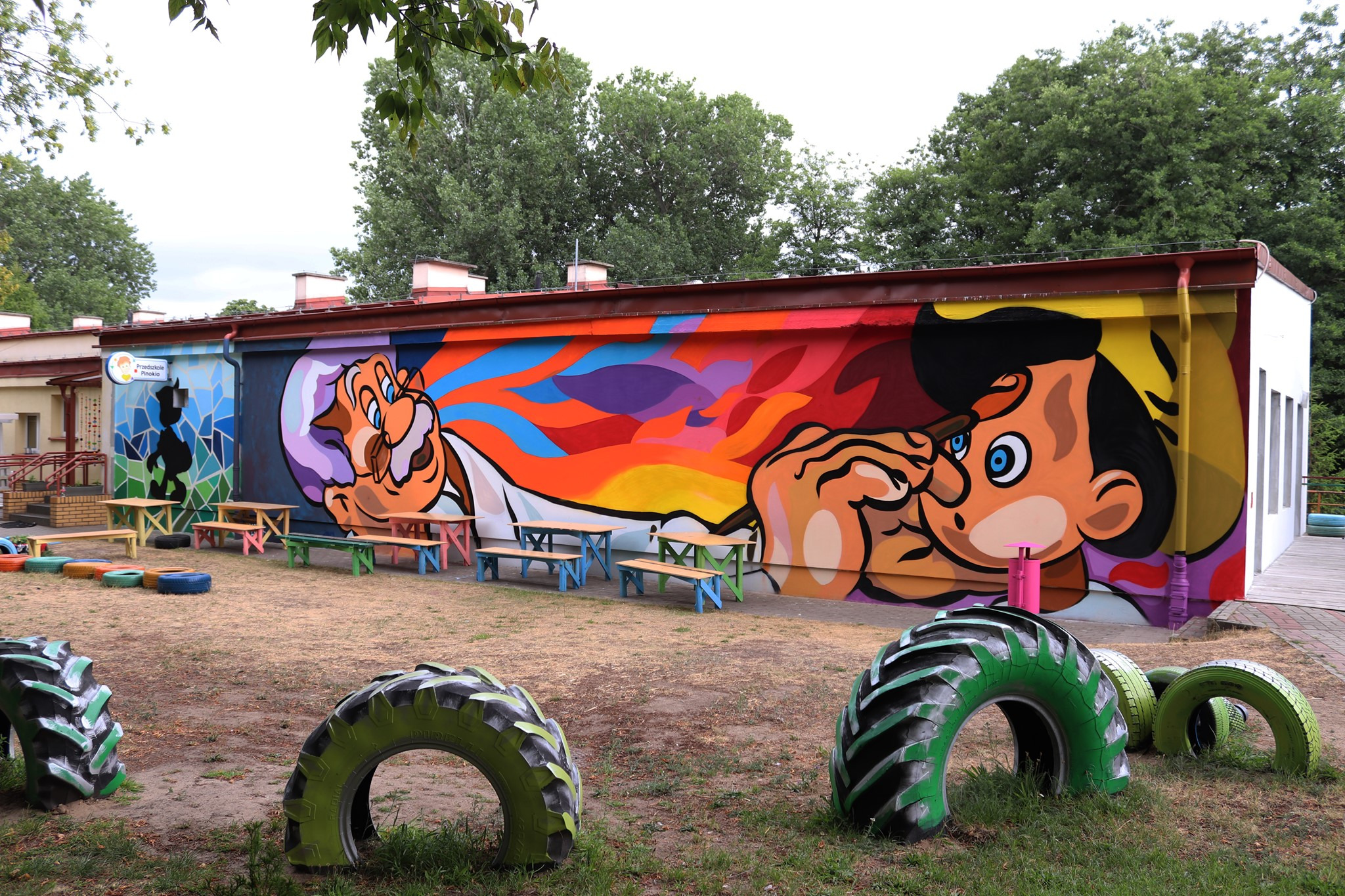 Kolorowa, bajkowa postać Pinokia na ścianie budynku przedszkola w Koninie, z oponami pomalowanymi na zielono w roli elementów placu zabaw.