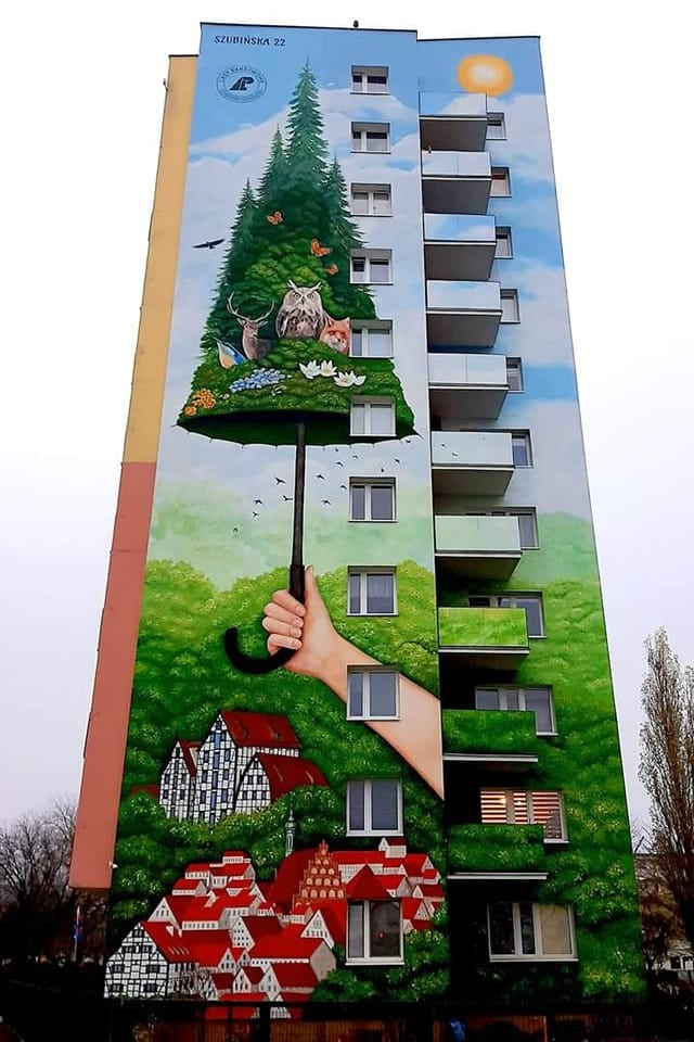 Wielkoformatowe malowidło na ścianie budynku przedstawiające rękę trzymającą parasol, pod którym znajduje się las z zwierzętami i tradycyjna zabudowa, słońce w prawym górnym rogu.