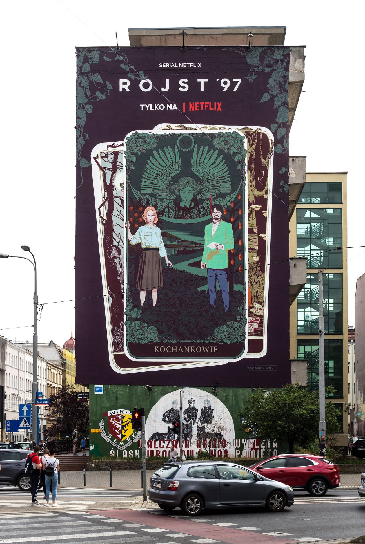 Wielkoformatowy mural na ścianie budynku przedstawiający grafikę inspirowaną kartami tarota i promujący serial Netflix 'Rojst '97', z widocznym fragmentem starego muralu poniżej, częściowo...