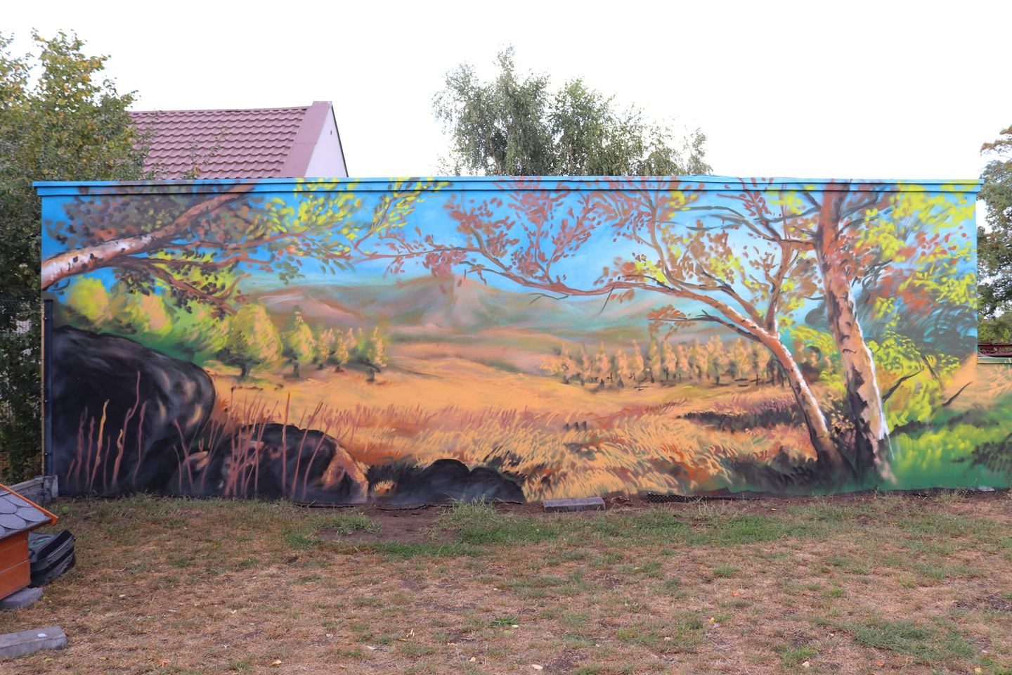 Krajobraz z drzewami i górami namalowany na metalowej ścianie, widoczne trawy i skały na pierwszym planie, obok trawnik i fragment dachu budynku.