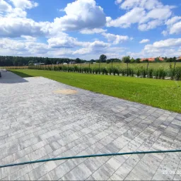 Widok na zadbany trawnik z systemem nawadniania, obok kostka brukowa i nowoczesny dom w słoneczny dzień.