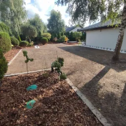 Przygotowany teren pod trawnik z rolki, obrzeża wyłożone kostką, widoczne młode drzewka i krzewy, w tle osoba pracująca przy domu.