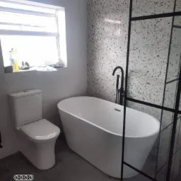 Nowoczesna łazienka z wolnostojącą białą wanną, czarną armaturą i kabiną prysznicową z czarnymi profilami, widok na toaletę i okno z środkami czystości.