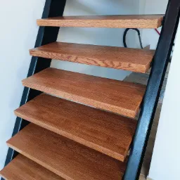 Nowoczesne schody wewnętrzne z drewnianymi stopniami o ciepłym odcieniu brązu i czarną metalową konstrukcją nośną, widoczne z dołu, z fragmentem podłogi wyłożonej płytkami imitującymi drewno.