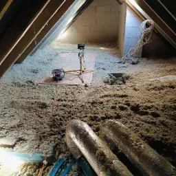 Poddasze w trakcie ocieplania, widoczne wełna mineralna, przewody wentylacyjne w srebrnej folii, niebieskie karbowane rury oraz lampy oświetleniowe na statywach.