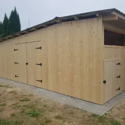 Nowo wybudowana drewniana konstrukcja z trzema parami drzwi na zawiasach i małym okienkiem, z widoczną konstrukcją dachu i elementami wykończeniowymi, otoczona zielenią i kostką brukową.