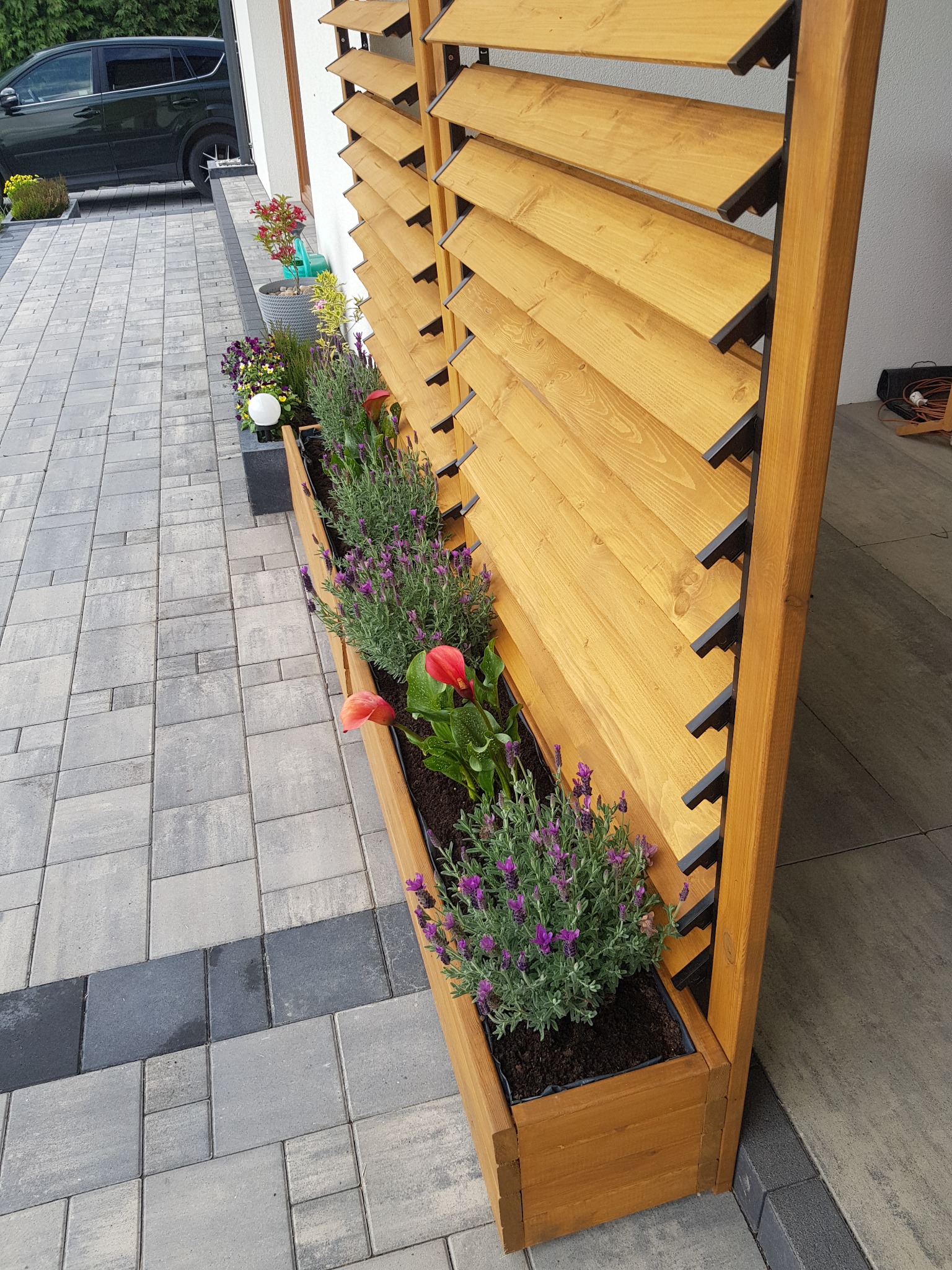 Drewniana konstrukcja z żaluzjami i wbudowanymi donicami z lawendą i anturium, ustawiona na wybrukowanym podjeździe, z widocznym samochodem w tle.