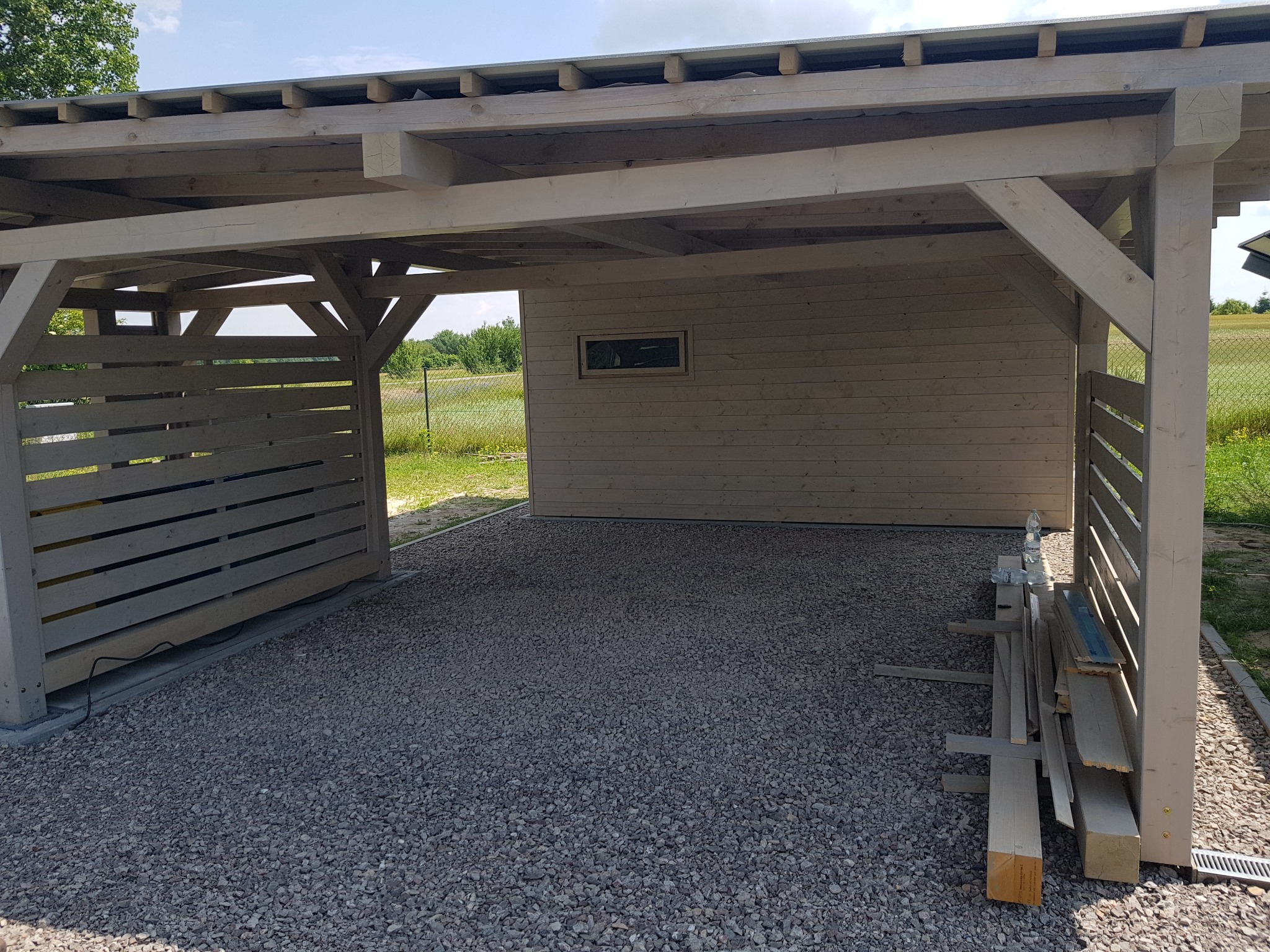 Drewniana altana z zadaszeniem w Ciecierzynie, z ażurową ścianą boczną i budynkiem gospodarczym w tle, na żwirowym podłożu. Przy ścianie altany leżą deski i butelka wody.